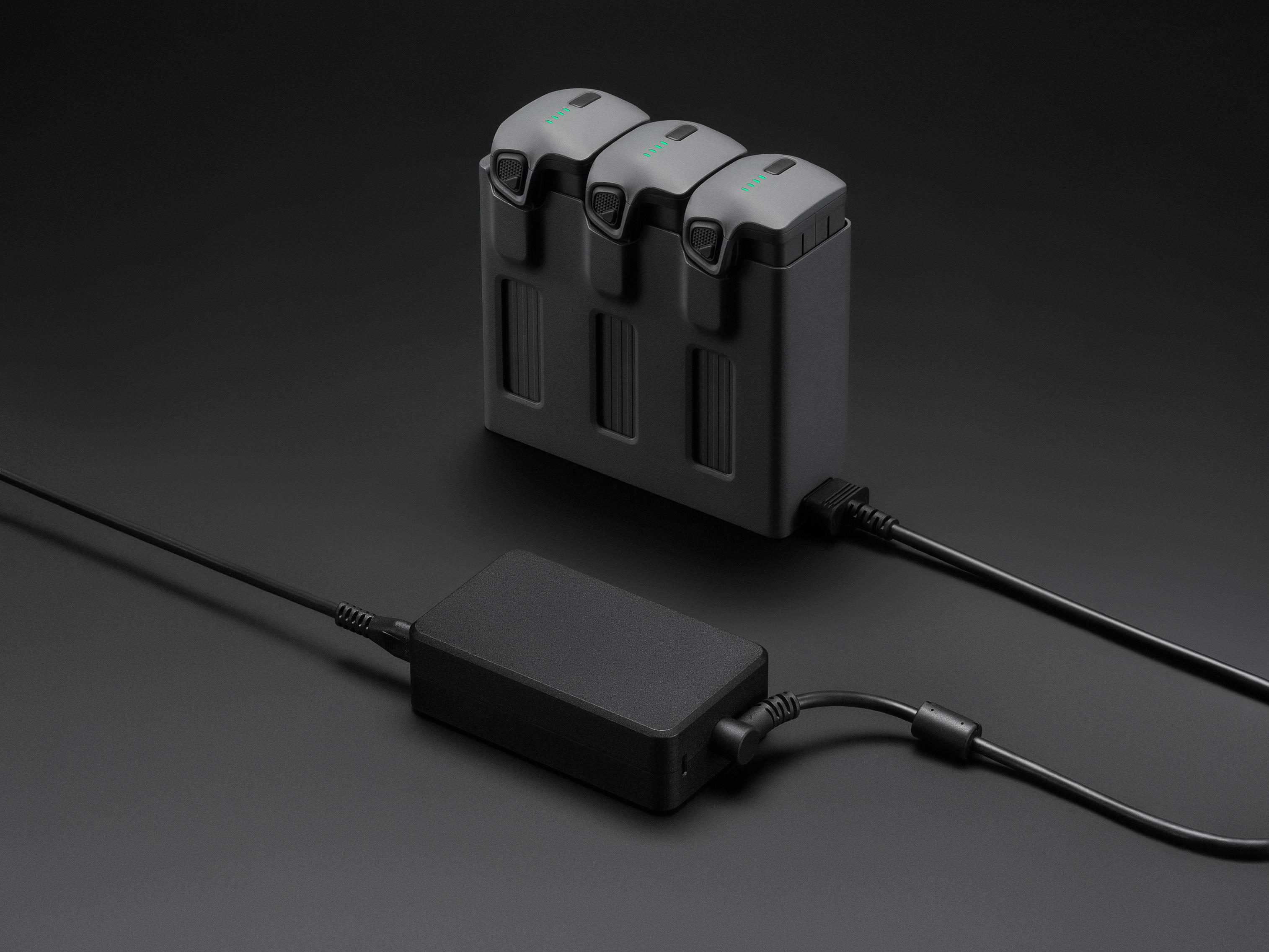 Eine Ladestation mit drei Drohnenbatterien und einem schwarzen Netzteil auf dunkler Oberfläche.