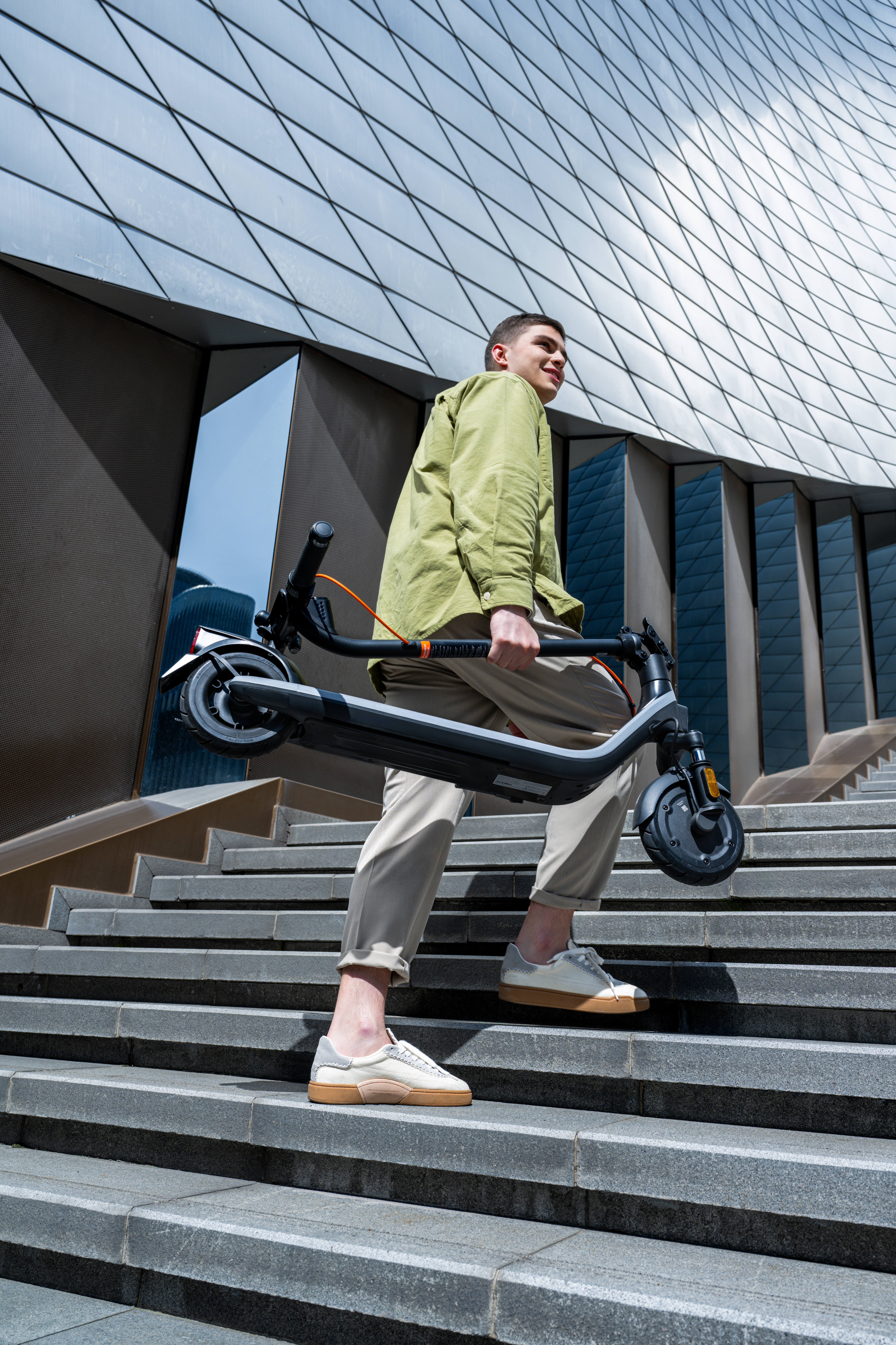 Bir adam siyah bir elektrikli scooter'ı merdivenlerden yukarı taşıyor. Arka planda modern bir bina var.