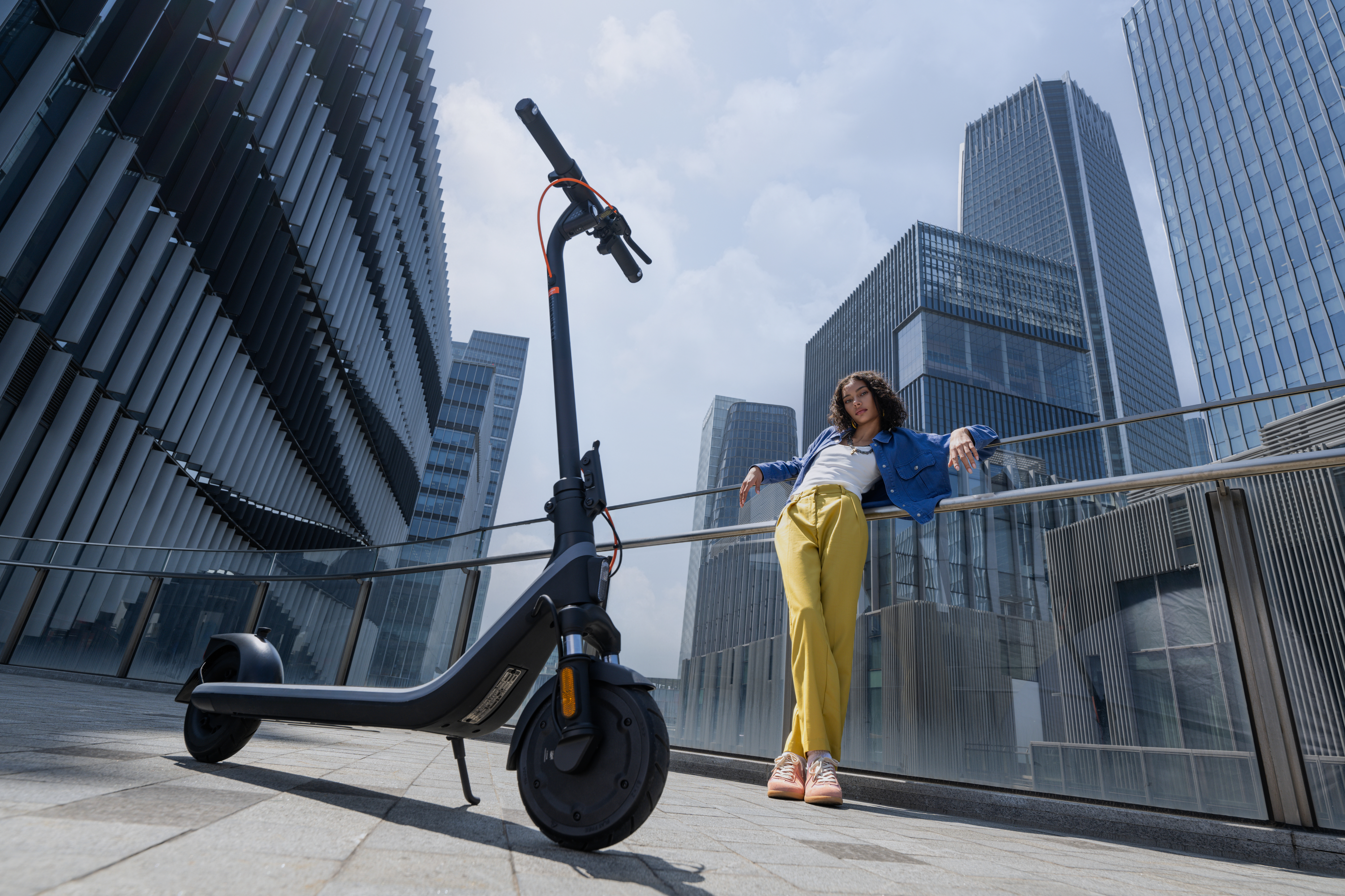 Bir kadın, bir scooter'ın yanında bir korkuluğa yaslanıyor. Bir adam scooter'ın yanında duruyor. Arka planda yüksek binalar.