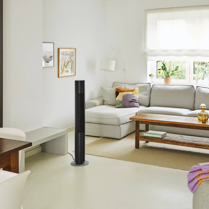 Czarny wentylator wieżowy w jasnym pokoju. Jest sofa, stół i okno.