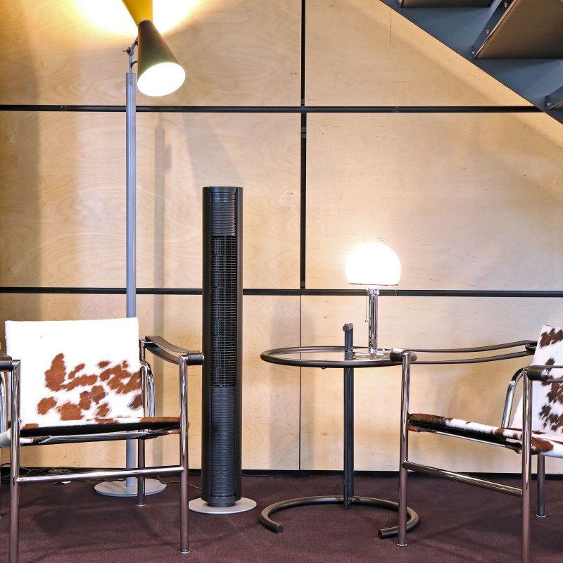 Czarny wentylator wieżowy w pokoju z dwoma krzesłami, stołem, lampą i drewnianymi panelami.