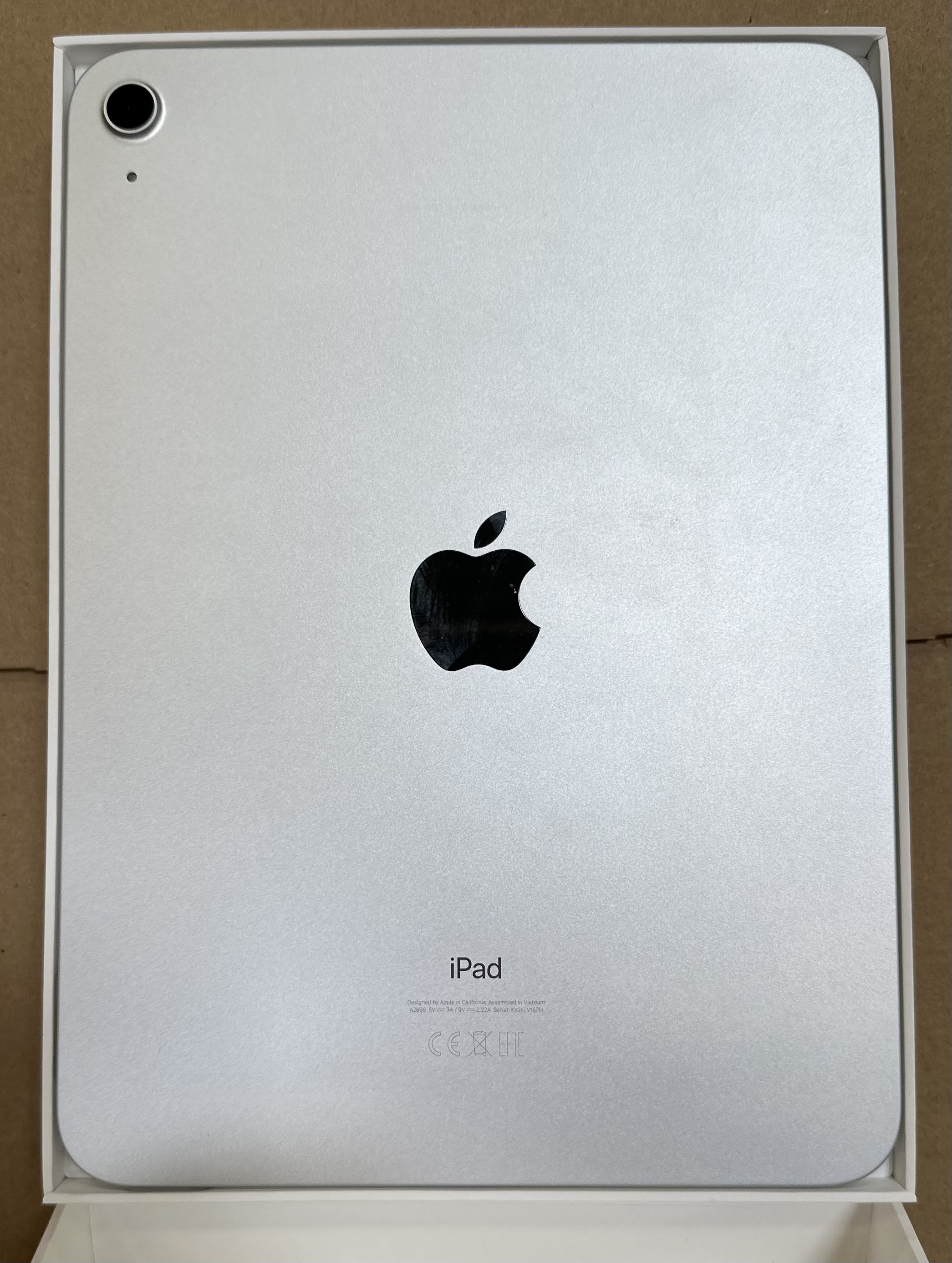 Tył srebrnego iPada w białym pudełku. Ma czarne logo Apple i aparat.