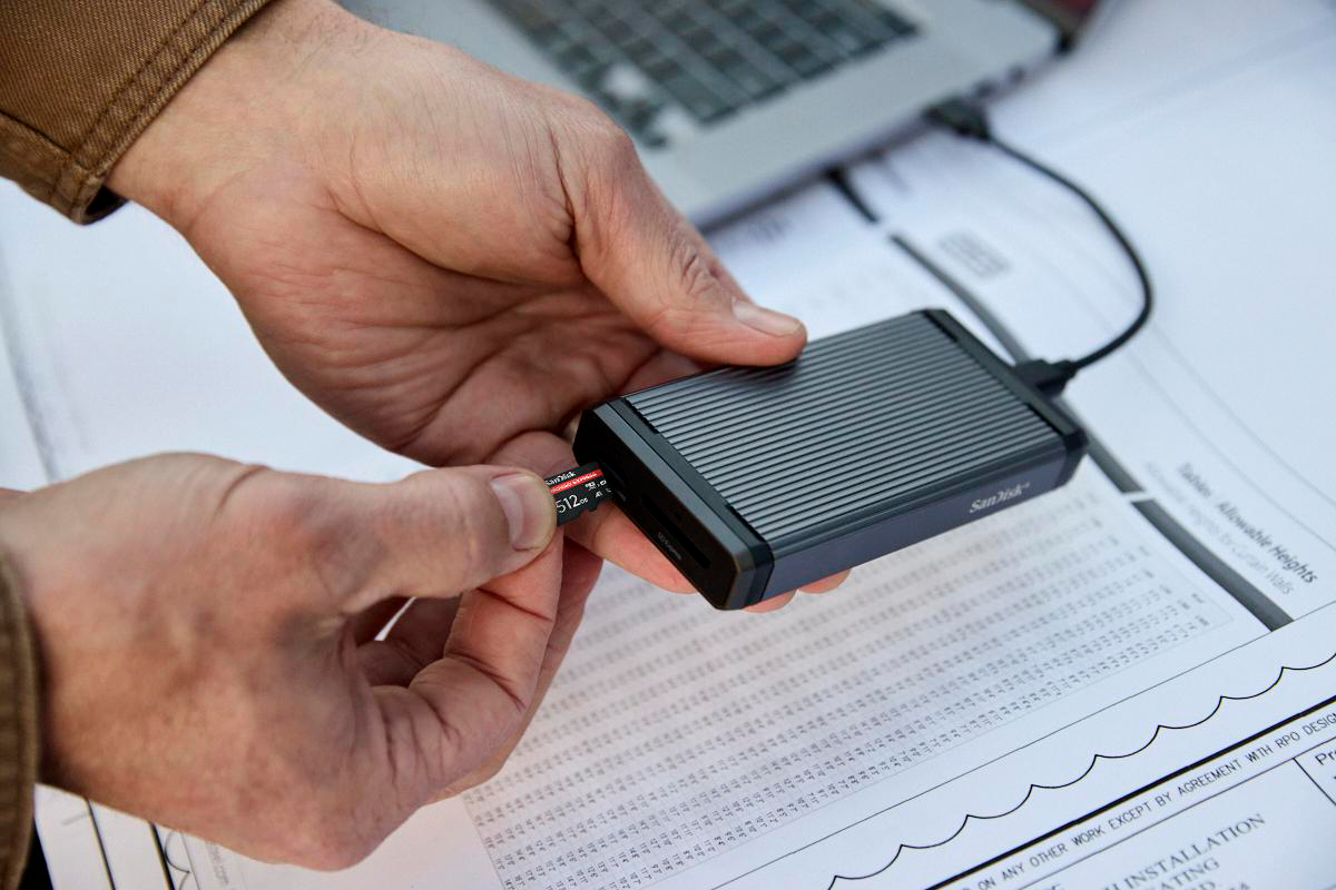 Hände stecken eine SanDisk microSD-Karte in ein Lesegerät. Baupläne und ein Laptop sind im Bild.