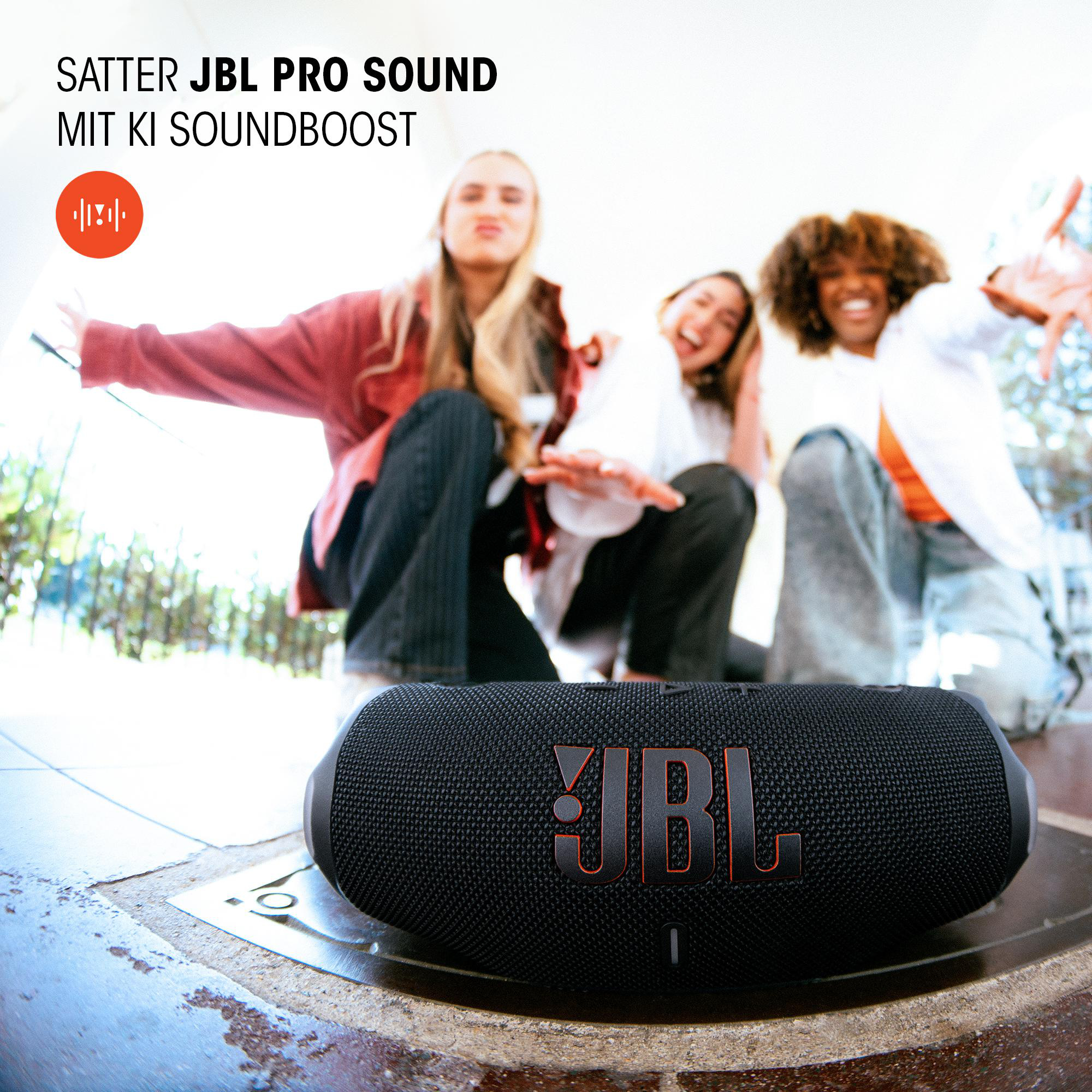Drei lächelnde Frauen mit einem schwarzen JBL-Lautsprecher auf einer Oberfläche.