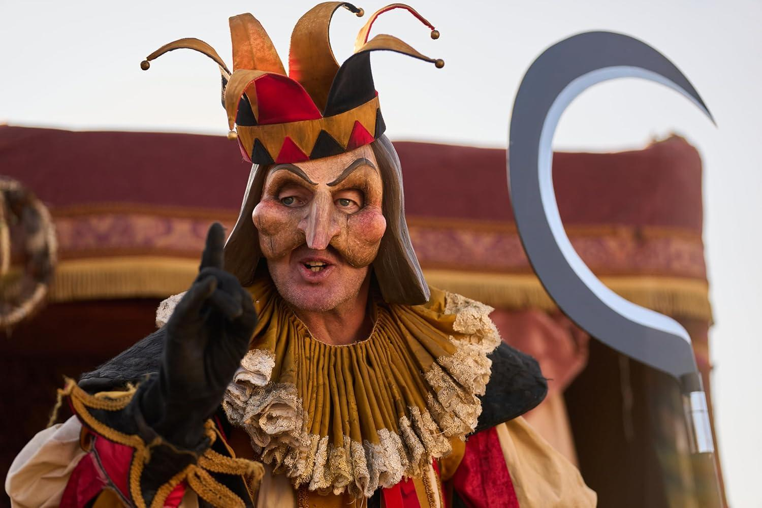 Ein Mann mit dramatischer Maske und einer Sichel. Er zeigt mit einem Finger. Roter und goldener Hintergrund.