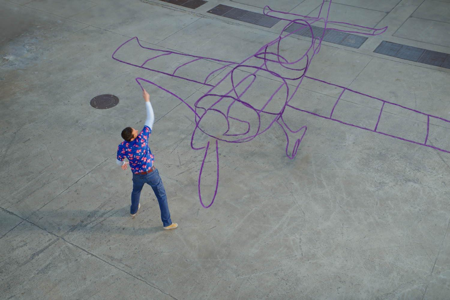 Mann zeichnet mit Kreide einen Flugzeugumriss auf Beton. Von oben gesehen. Blaues Hemd, Jeans.
