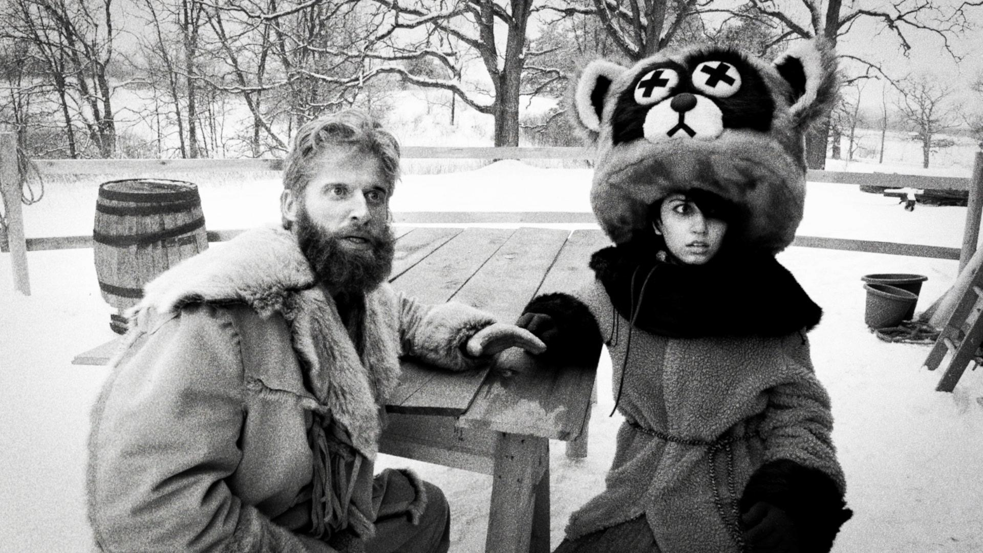 Ein Mann mit Bart und eine Frau mit Waschbärhut sitzen im Schnee an einem Tisch.