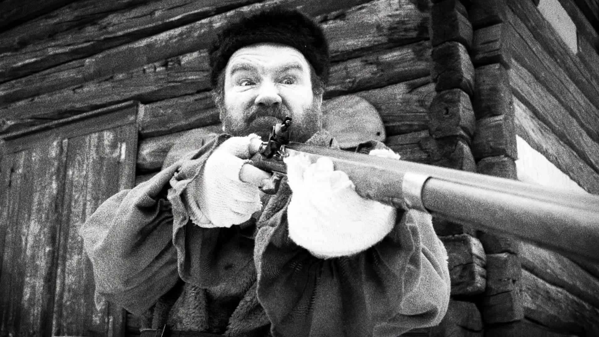 Ein Mann mit Bart zielt mit einem Gewehr, im Hintergrund ein Holzgebäude.