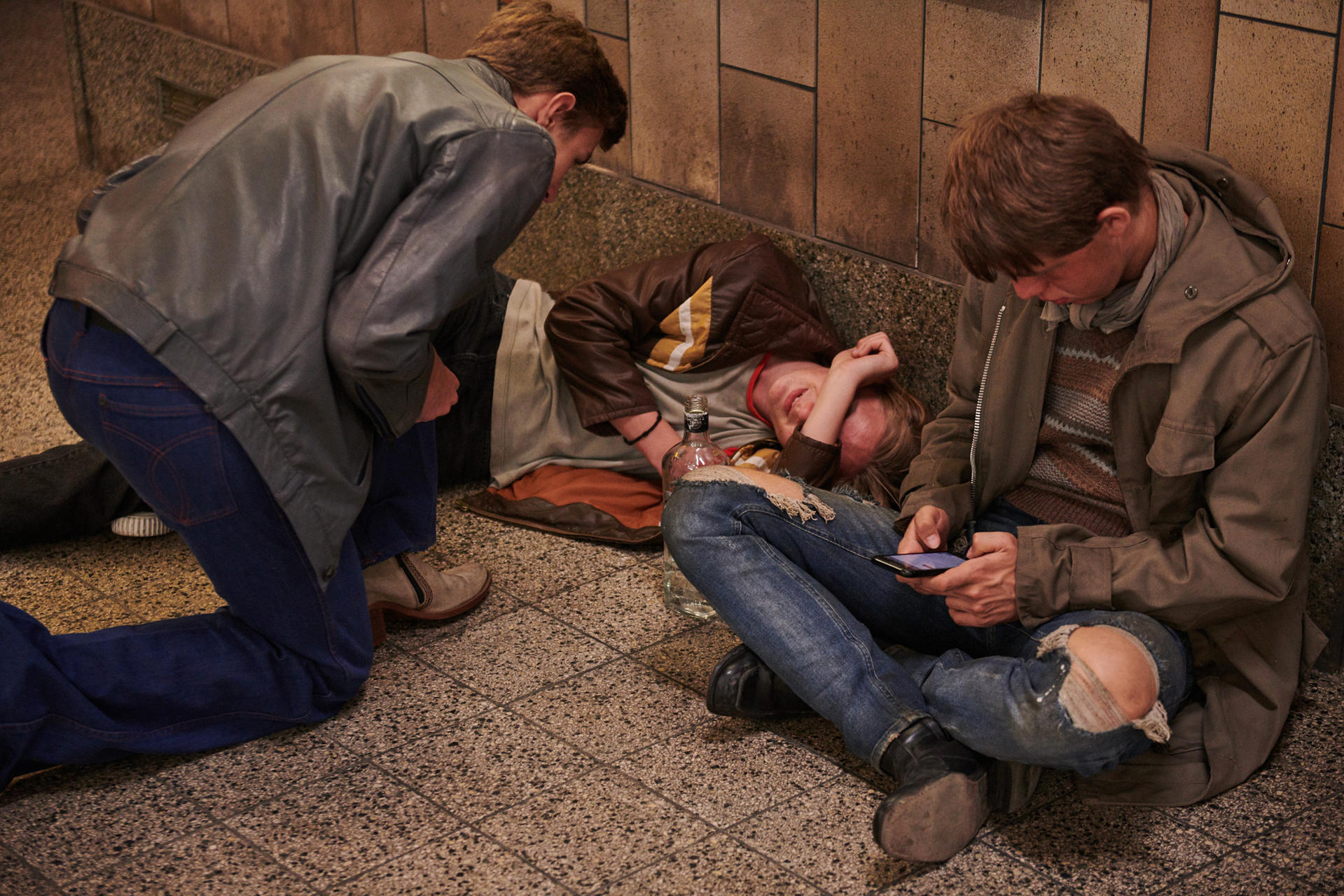 Drei Männer sitzen in einer U-Bahn. Einer schläft, einer schaut auf ein Telefon. Der Boden ist gefliest.