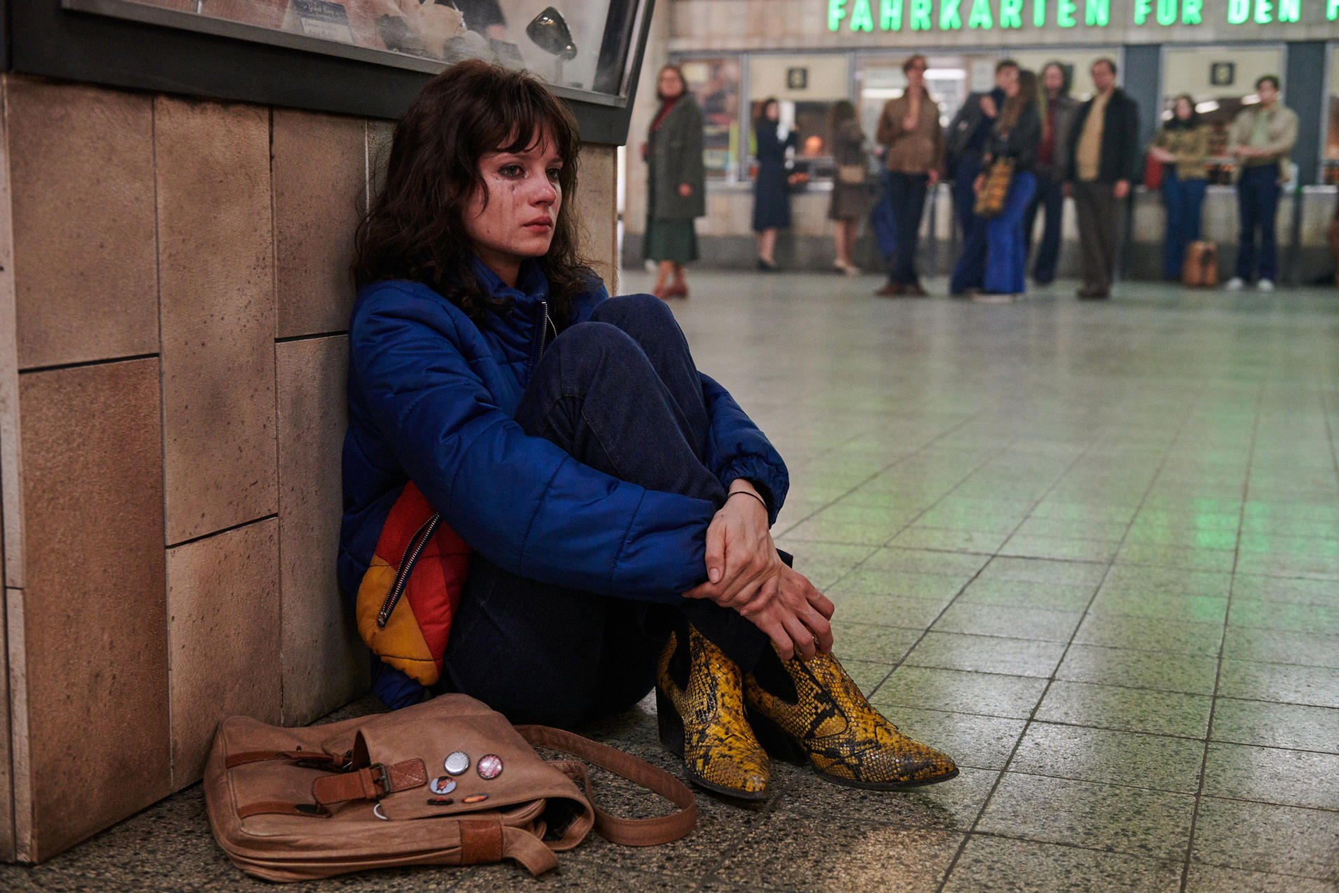 Eine Frau sitzt in einem Bahnhof. Sie weint. Auf dem Boden liegt eine Tasche.