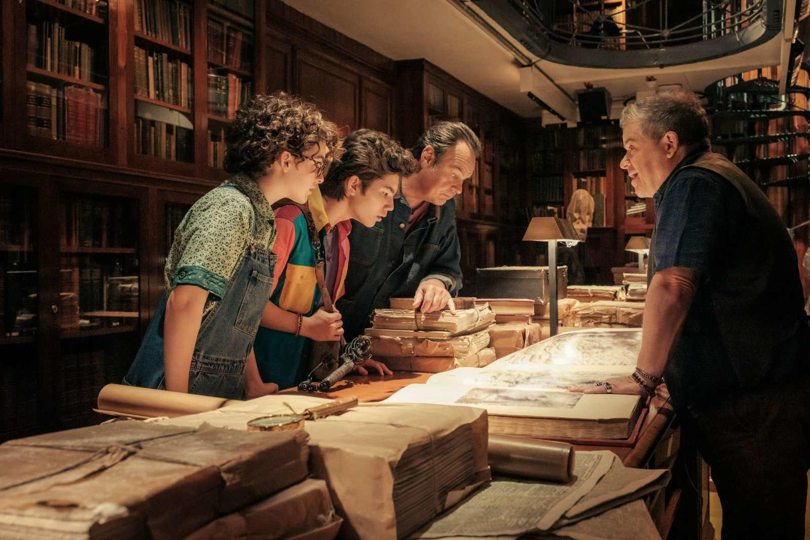 Vier Personen in einer Bibliothek, die sich alte Bücher und Karten ansehen. Bücherregale füllen den Hintergrund.