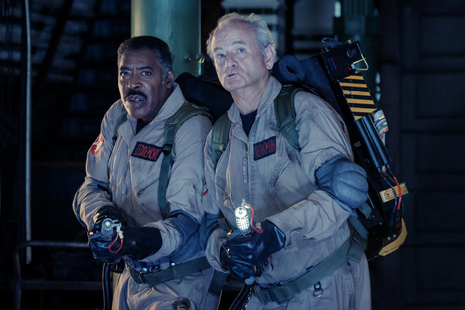 Zwei Ghostbusters in Overalls, halten Protonenpacks, sehen alarmiert aus. Dunkler Raum Hintergrund.