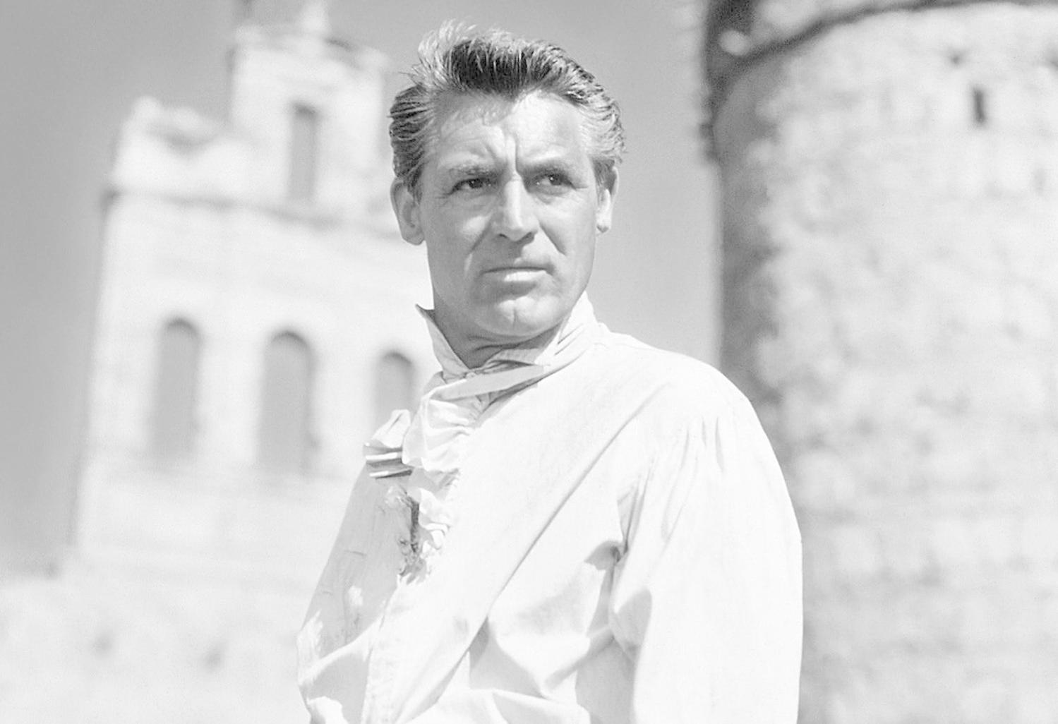 Cary Grant in Schwarzweiß, blickt weg, trägt ein weißes Hemd vor einer Burg.