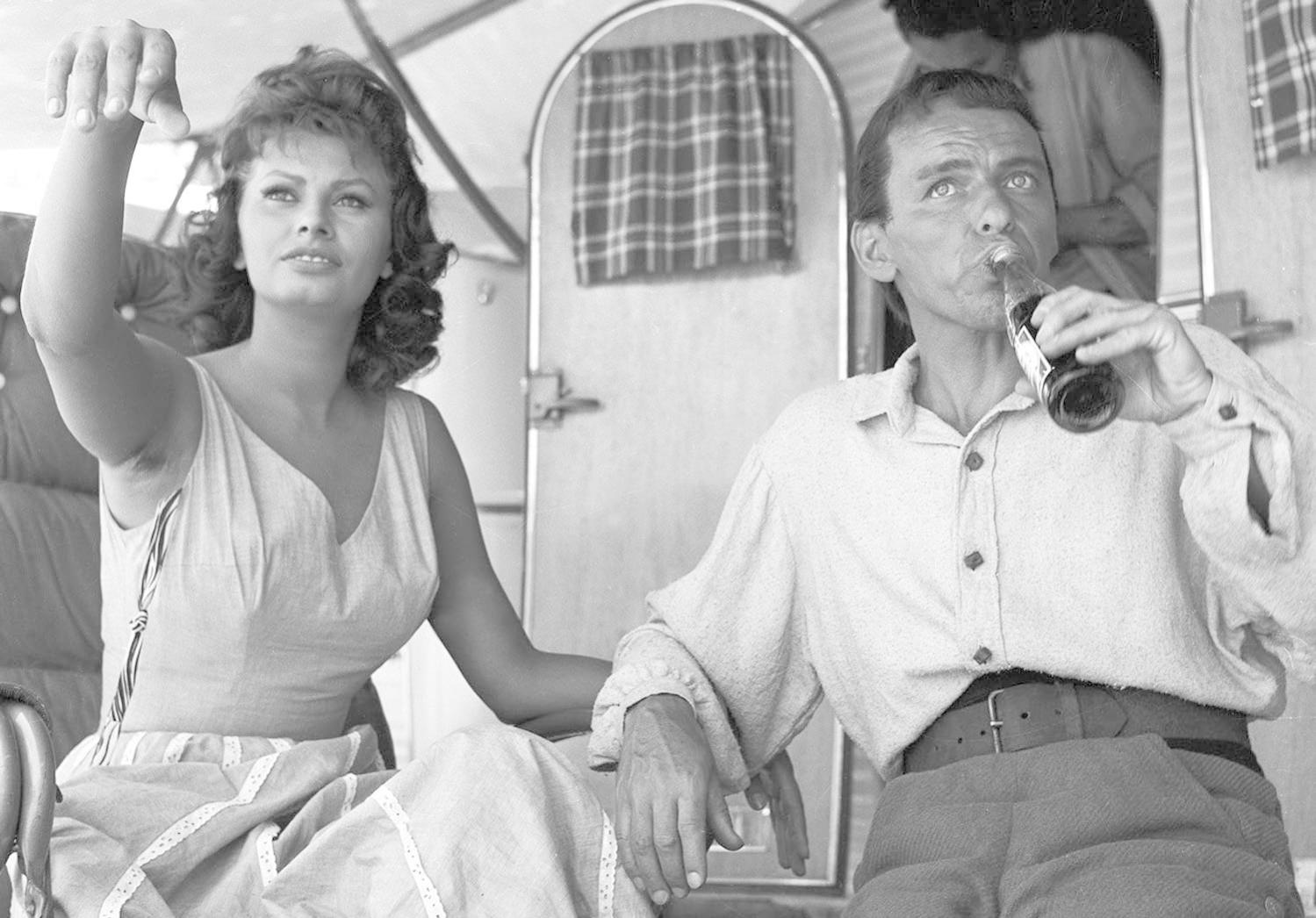 Sophia Loren und Frank Sinatra mit einer Flasche, in Schwarzweiß, im Freien.