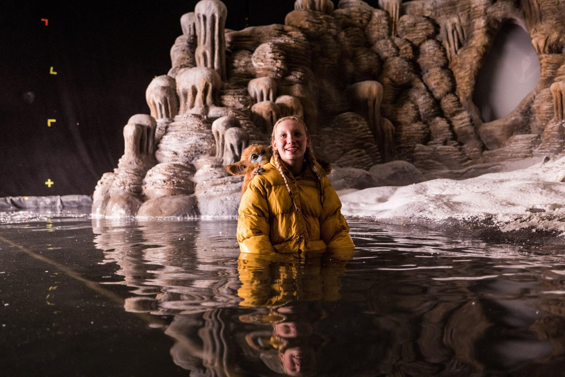 Ein Mädchen in gelber Jacke im Wasser mit einem Reh. Dahinter eine eisartige Struktur.