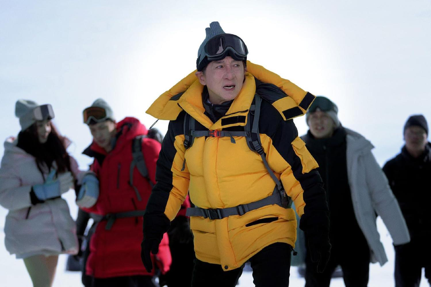 Jackie Chan in gelber Jacke und Brille, umgeben von Menschen in verschneiter Landschaft.