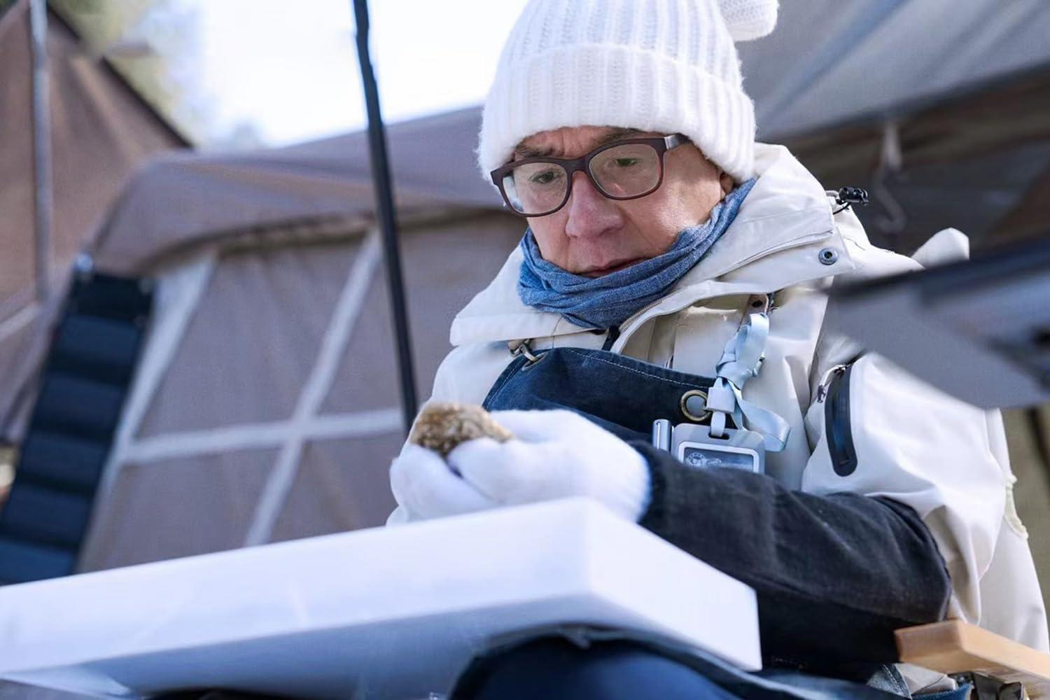Ein älterer Mann mit Brille und weißem Hut untersucht ein kleines Objekt. Er ist draußen.
