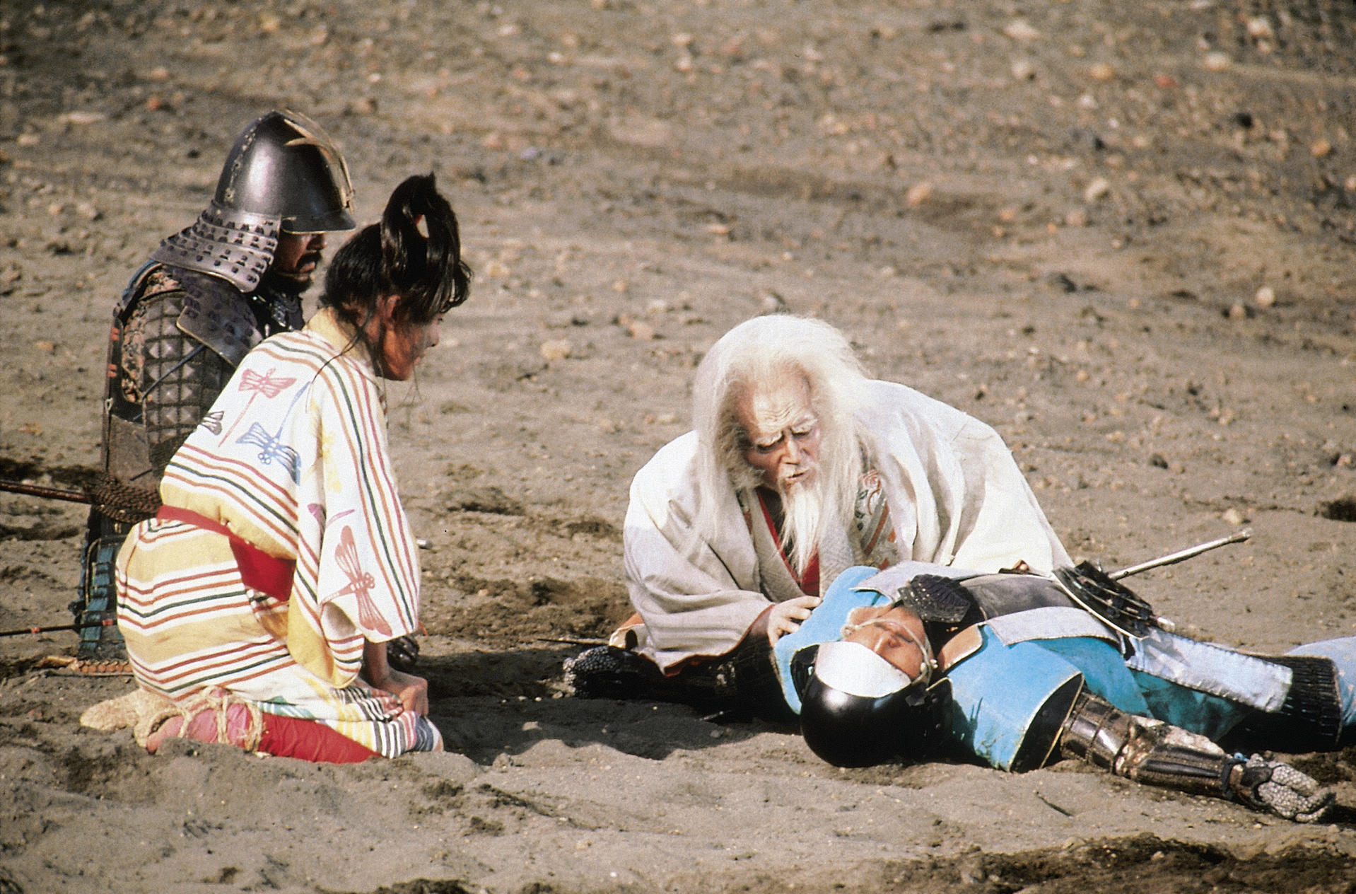 Ein Mann in Samurai-Rüstung liegt tot. Zwei Leute trauern um ihn. Szene an einem sandigen Ufer.