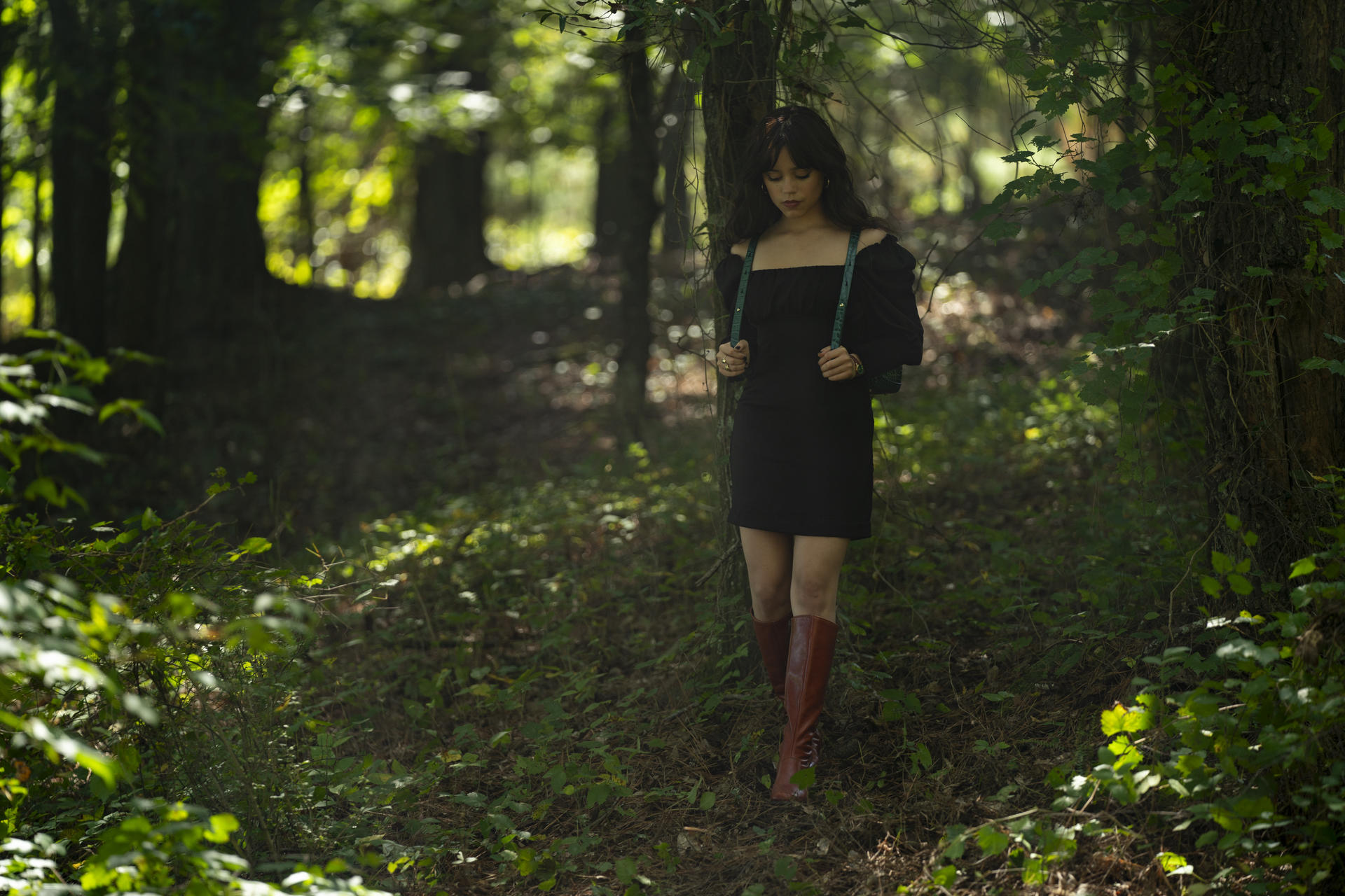 Frau in schwarzem Kleid geht durch einen Wald, Bäume im Hintergrund.