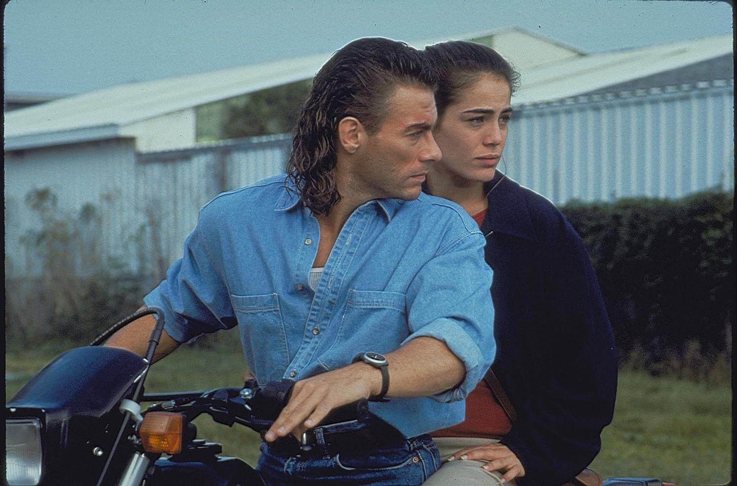 Van Damme auf einem Motorrad mit einer Frau hinter sich, die zur Seite blickt, Tageszeit-Szene.
