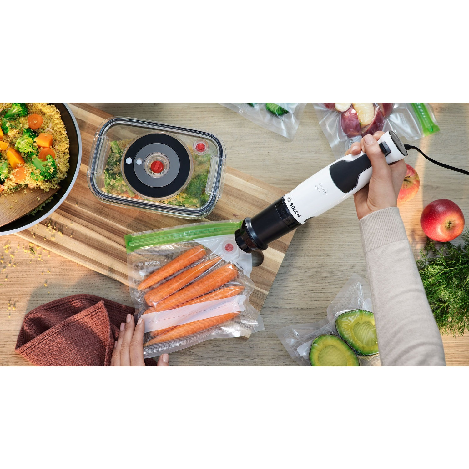 Una persona usa un frullatore Bosch per sigillare sottovuoto il cibo. Carote, broccoli e altri articoli visibili.