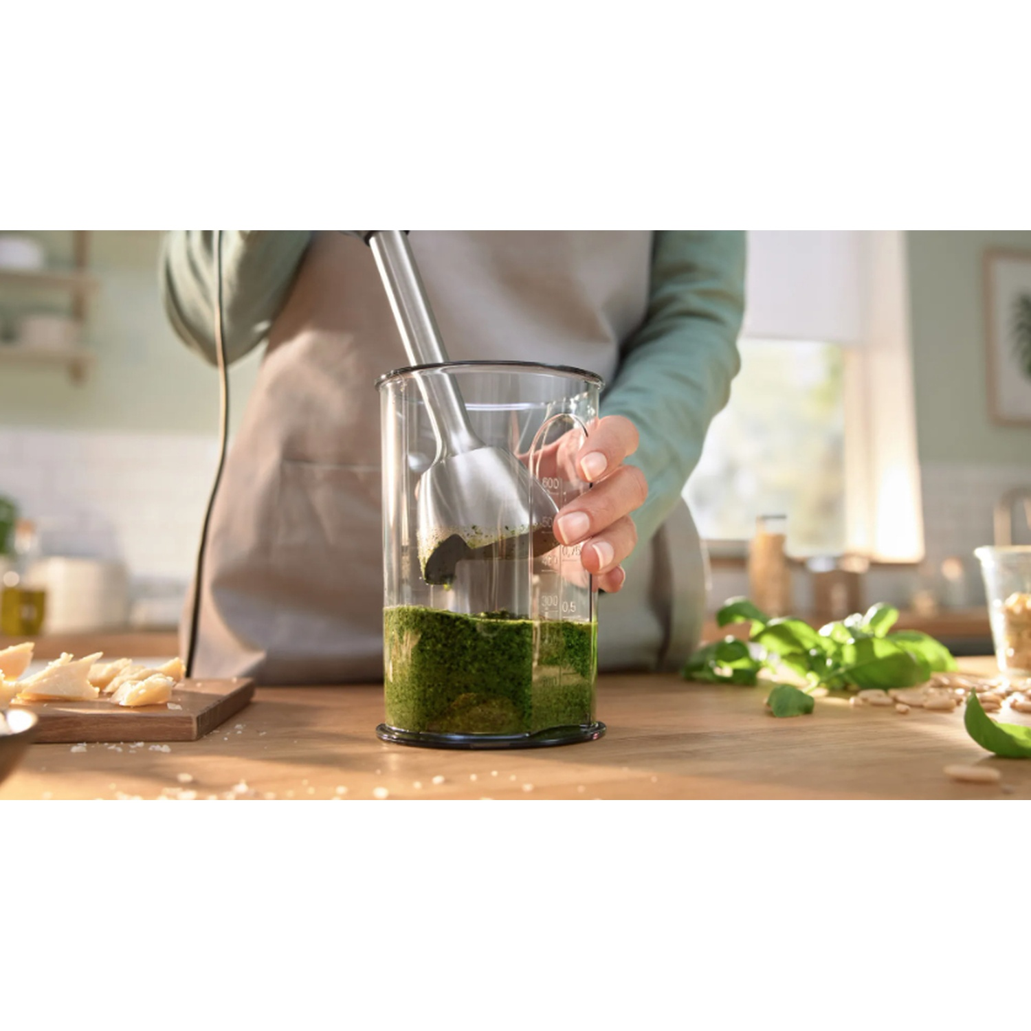 Una mano che frulla il pesto con un frullatore a immersione in un bicchiere. Altri ingredienti visibili.