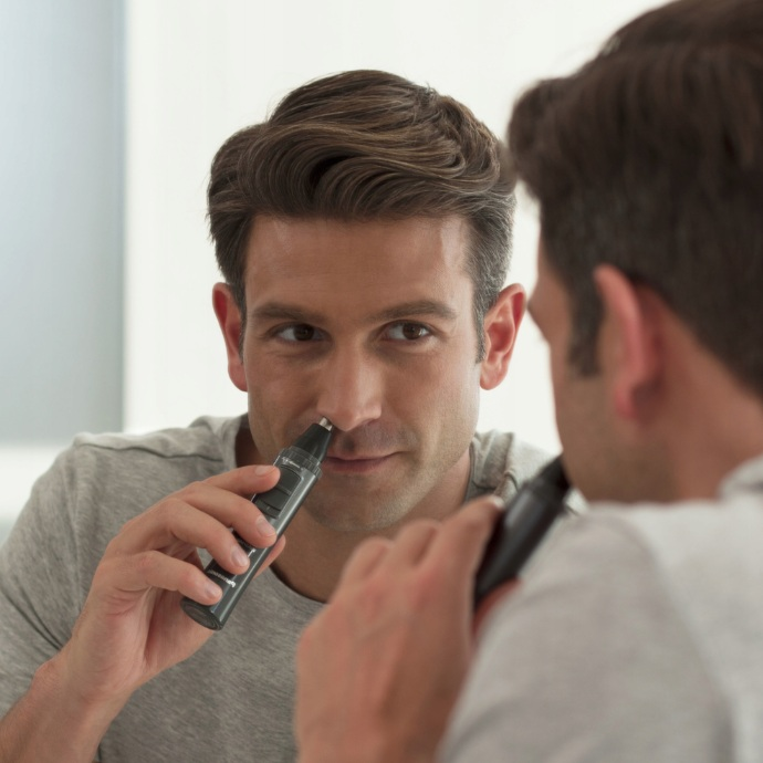 Mężczyzna używa trymera do nosa. Odbicie pokazuje tę samą czynność. Biała łazienka.