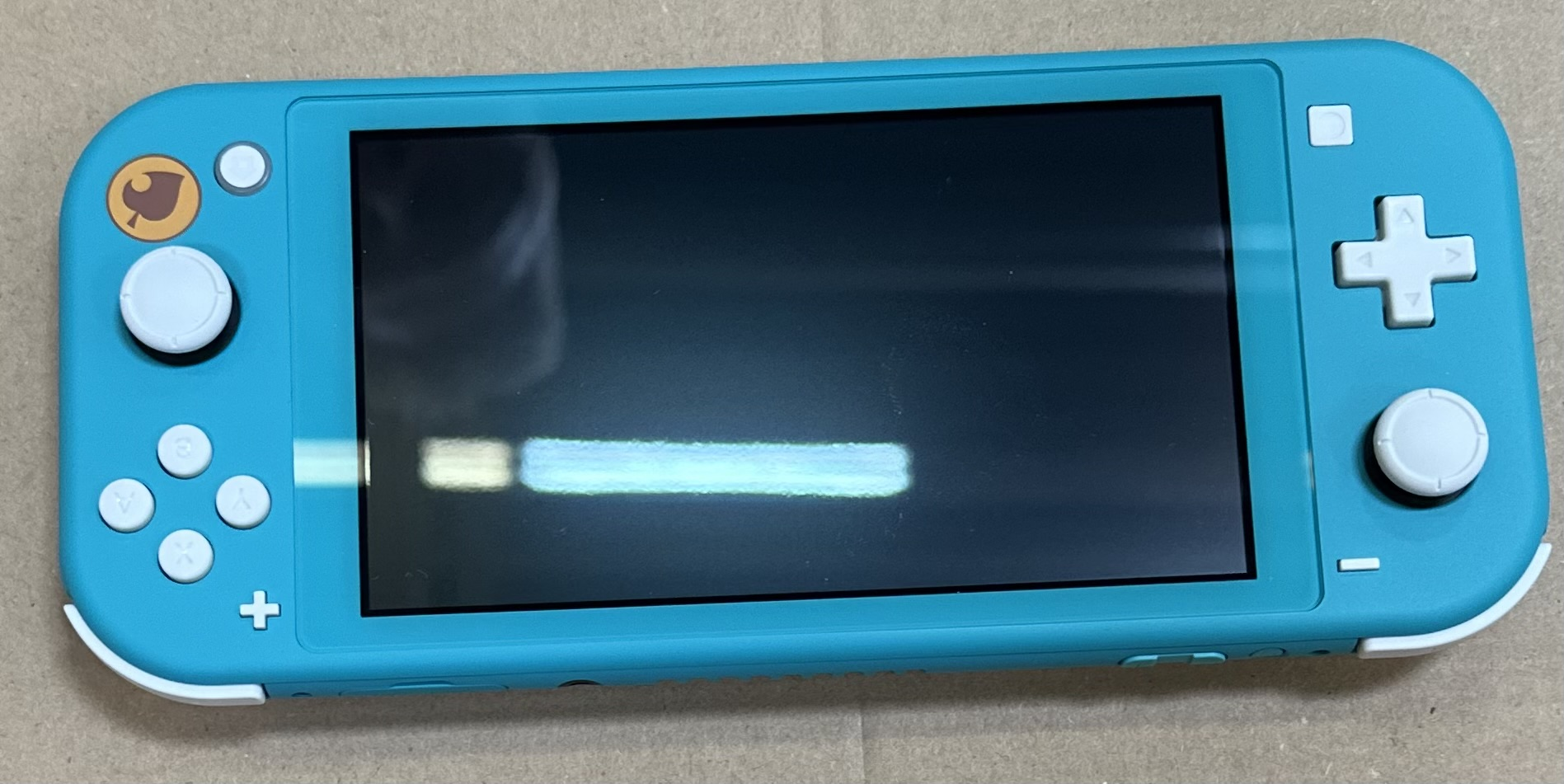 Konsola Nintendo Switch Lite w kolorze turkusowym. Ekran włączony, białe przyciski, brązowe tło.