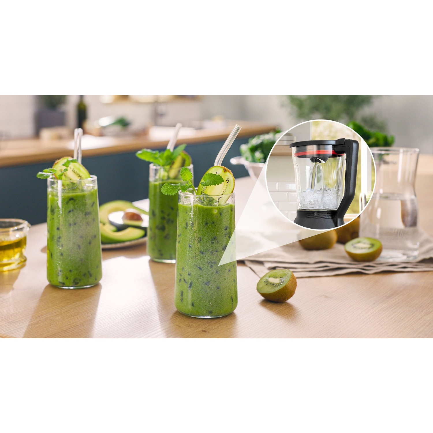 Tre frullati verdi in bicchieri, con guarnizione di kiwi e menta, accanto a un frullatore con ghiaccio dentro.