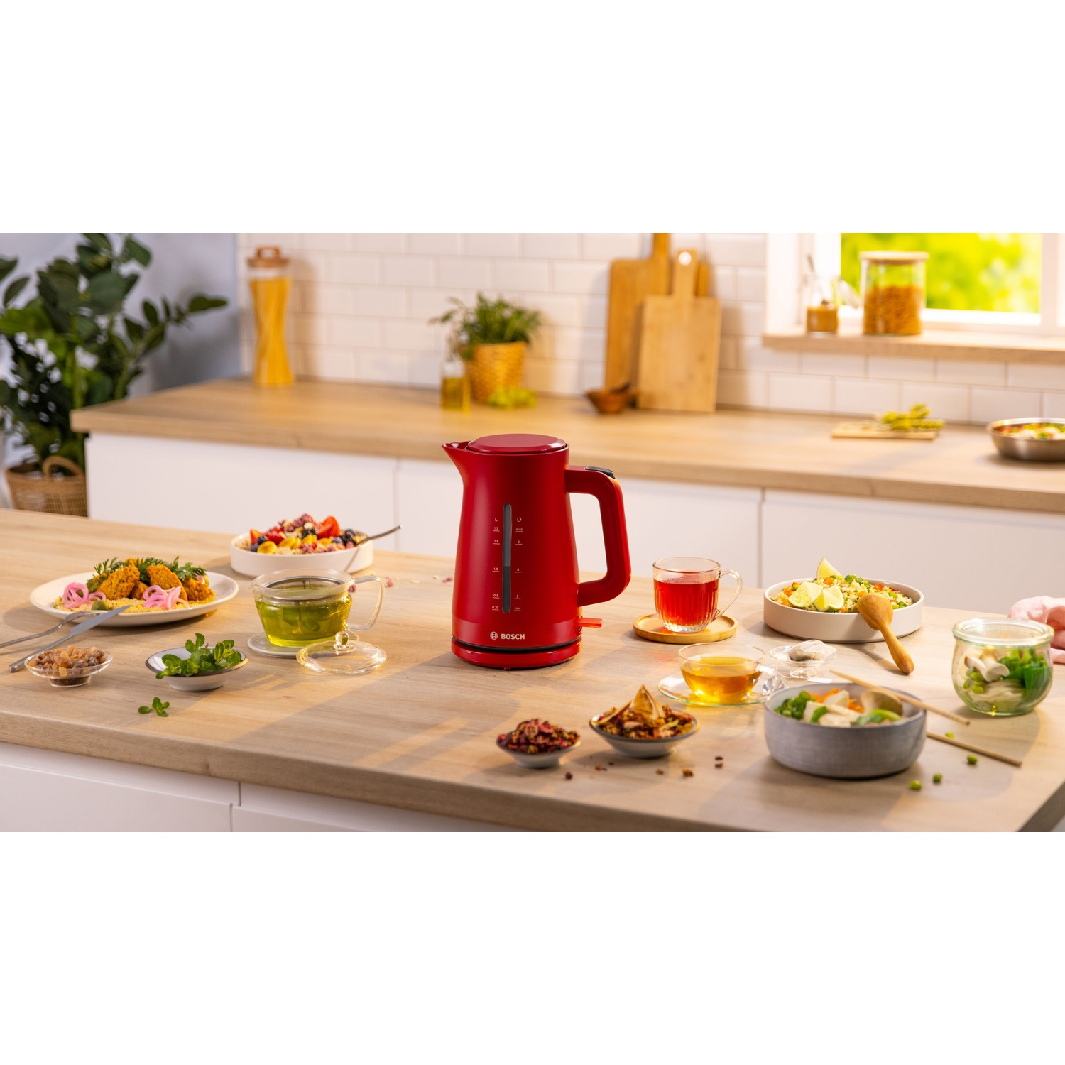 Un bollitore elettrico rosso su una superficie di legno con generi alimentari e altri utensili da cucina. Una finestra è sullo sfondo.