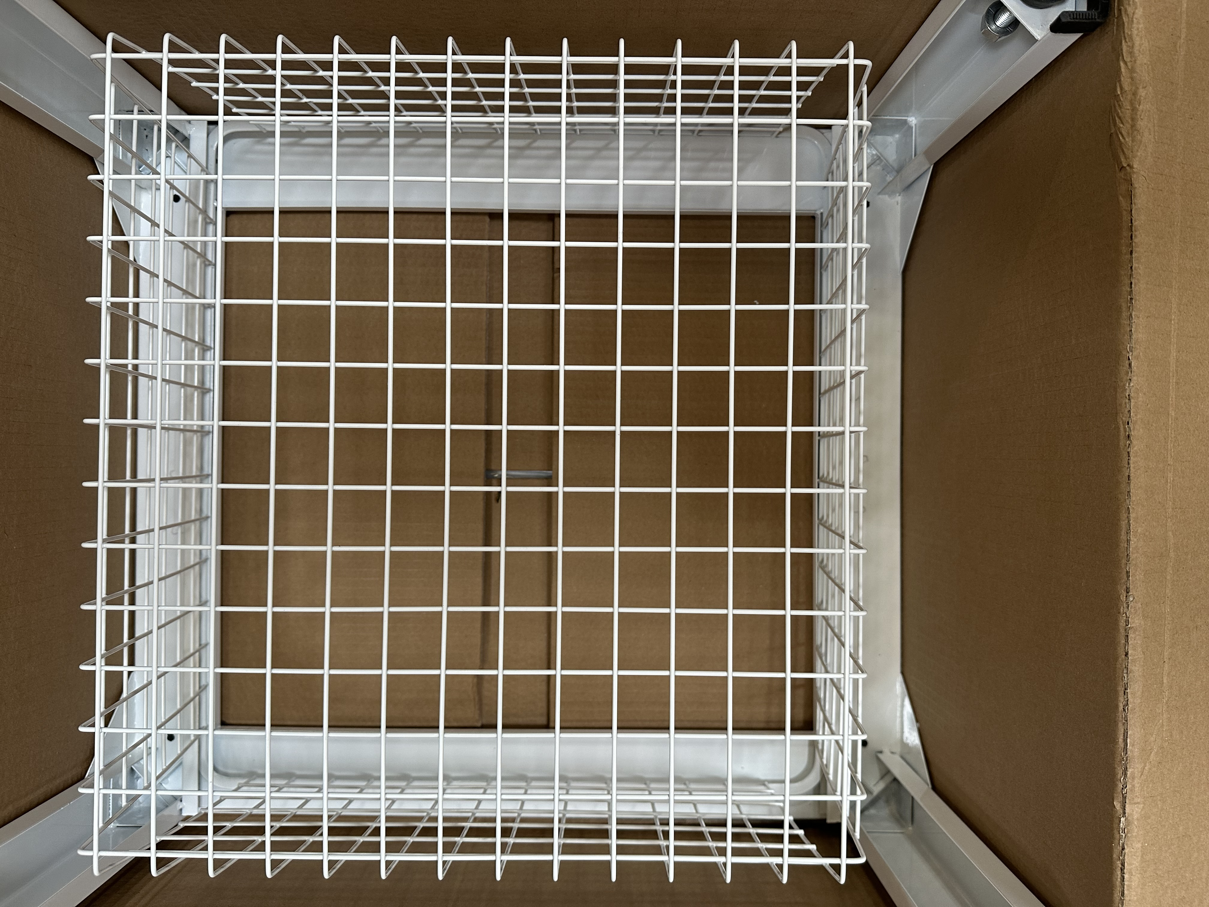 Biała metalowa półka druciana wewnątrz kartonowego pudełka, widok z góry.