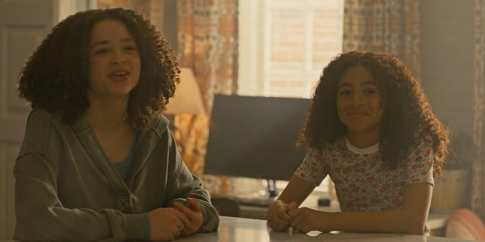 Zwei junge Mädchen mit lockigem Haar sitzen an einem Tisch. Eines spricht, das andere lächelt.