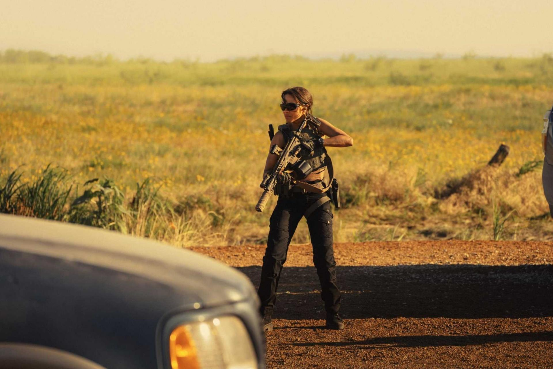 Frau mit Gewehr steht neben einem Auto auf einem Feld. Gelbe Blumen im Hintergrund an einem sonnigen Tag.