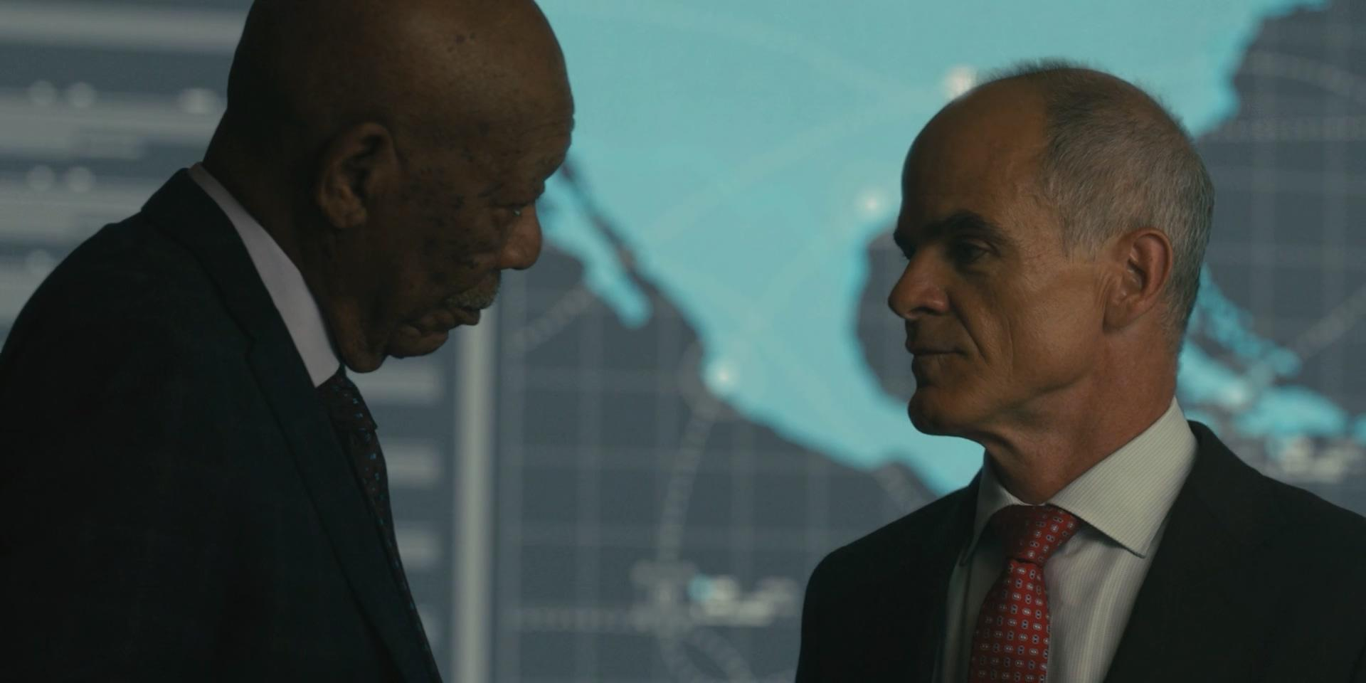 Zwei Männer in Anzügen stehen in einem Büro. Einer ist älter, schaut nach unten, der andere schaut ihn an.