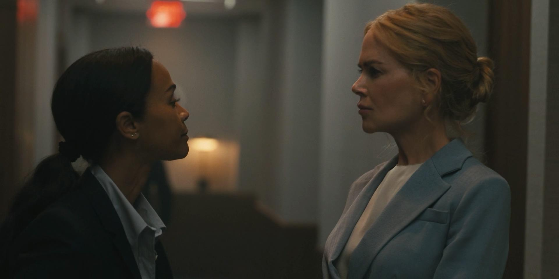 Zwei Frauen stehen sich in einem Flur gegenüber. Eine trägt einen Anzug, die andere einen hellblauen Mantel.