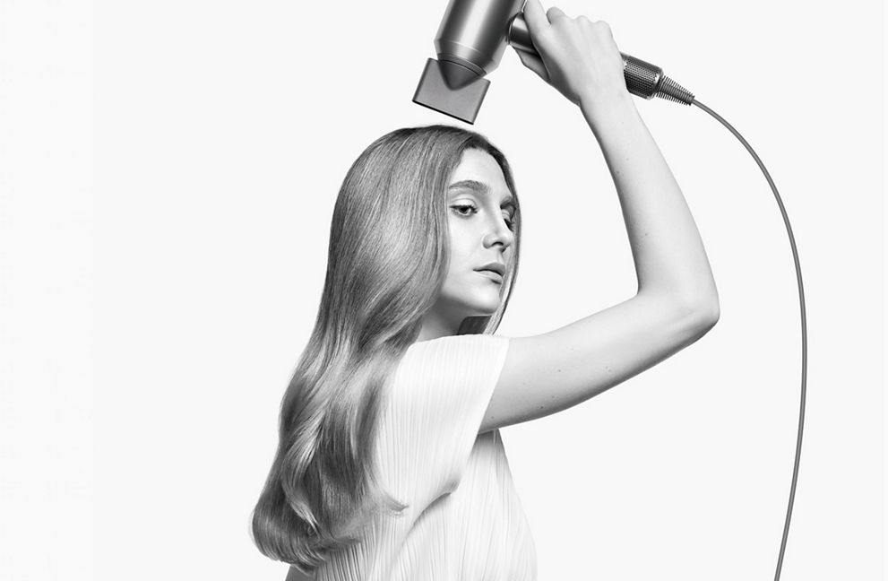 Une femme se sèche les longs cheveux avec un sèche-cheveux Dyson, en noir et blanc.