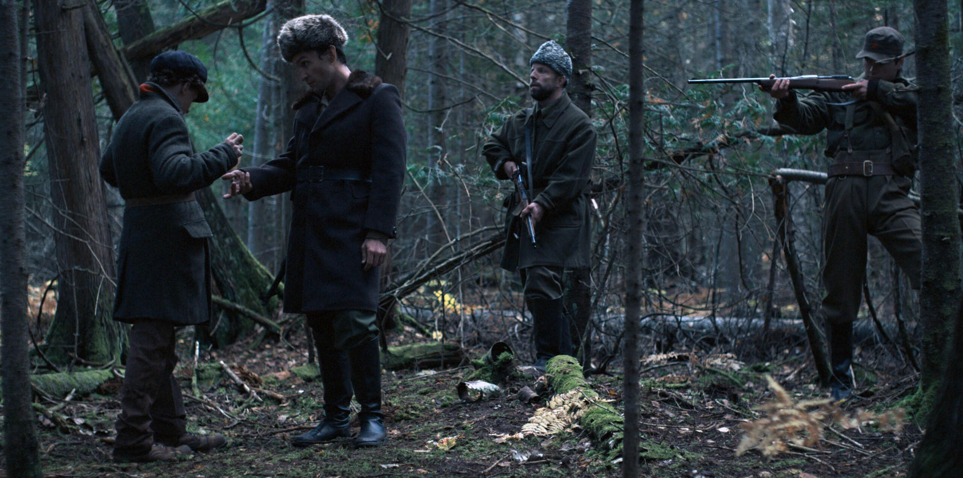 Männer in Mänteln im Wald, einige mit Gewehren; eine Diskussion findet statt.