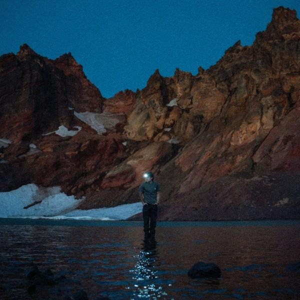 Eine Person steht im Wasser bei Dämmerung, trägt eine Stirnlampe, Berge im Hintergrund.