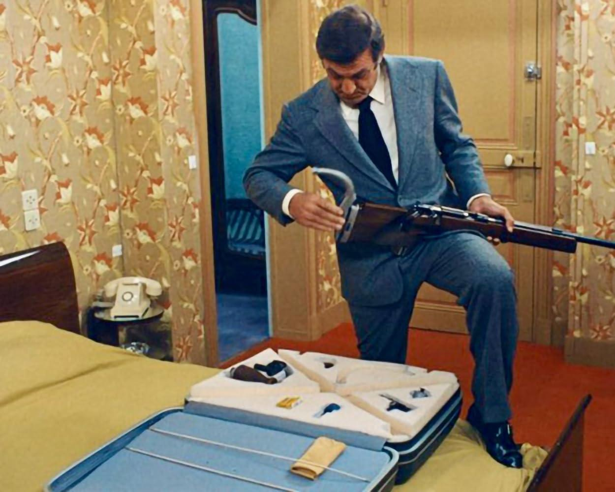 Ein Mann im Anzug untersucht ein Gewehr in einem geöffneten Koffer in einem Hotelzimmer mit Bett.