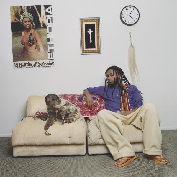 Mann mit Dreadlocks sitzt mit Hund auf Sofa, Poster und Uhr an der Wand.