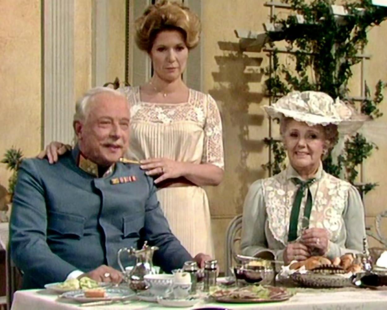 Drei Personen an einem Tisch. Ein älterer Mann, eine Frau und eine andere Frau dahinter. Auf dem Tisch steht Essen.