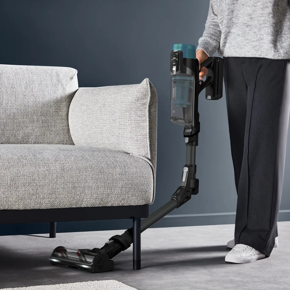 Osoba odkurzająca odkurzaczem pionowym. Szara kanapa po lewej. Ciemnoniebieska ściana z tyłu.
