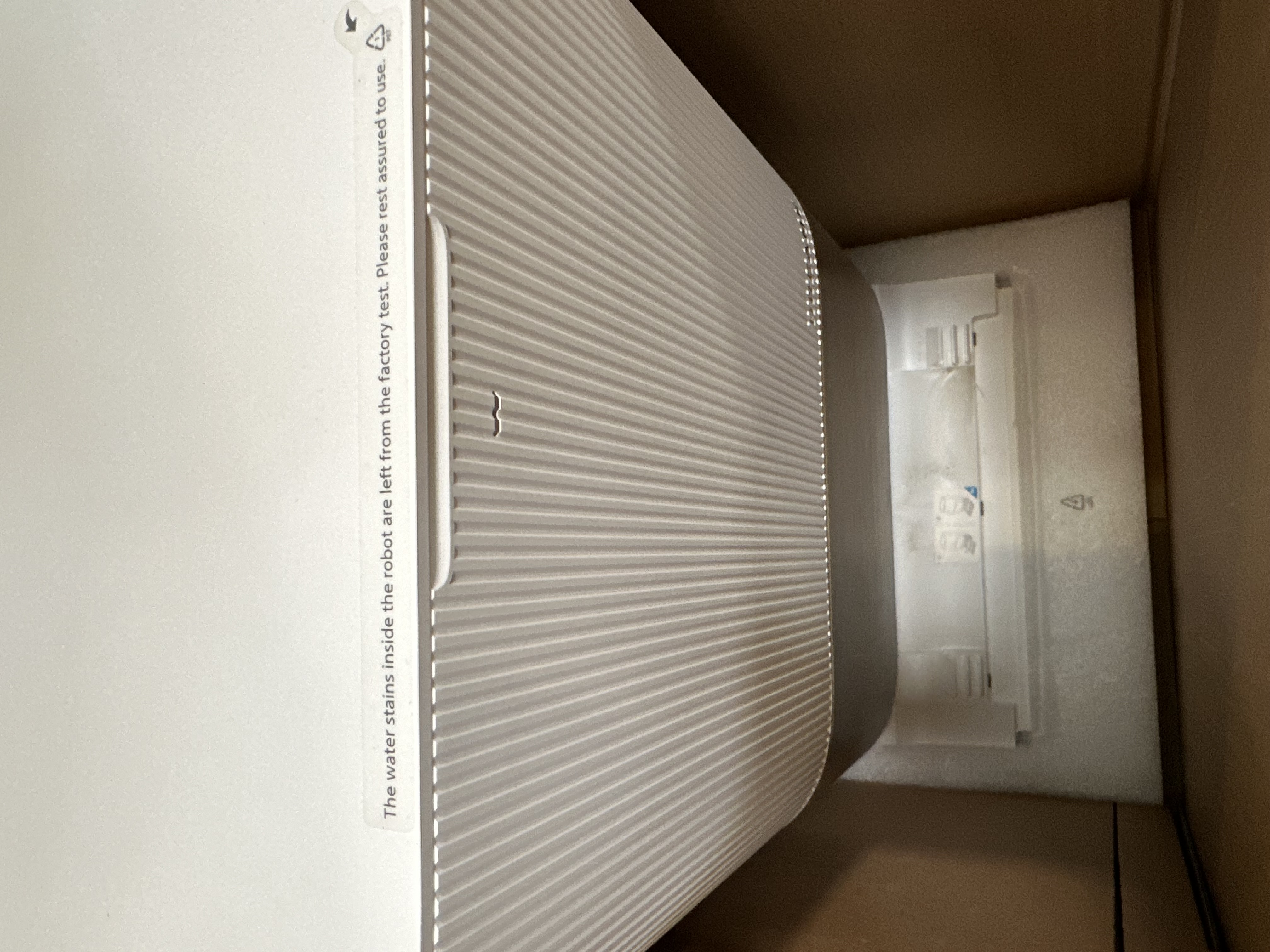 Biały odkurzacz robot wewnątrz kartonowego pudełka, z tekstem z boku.