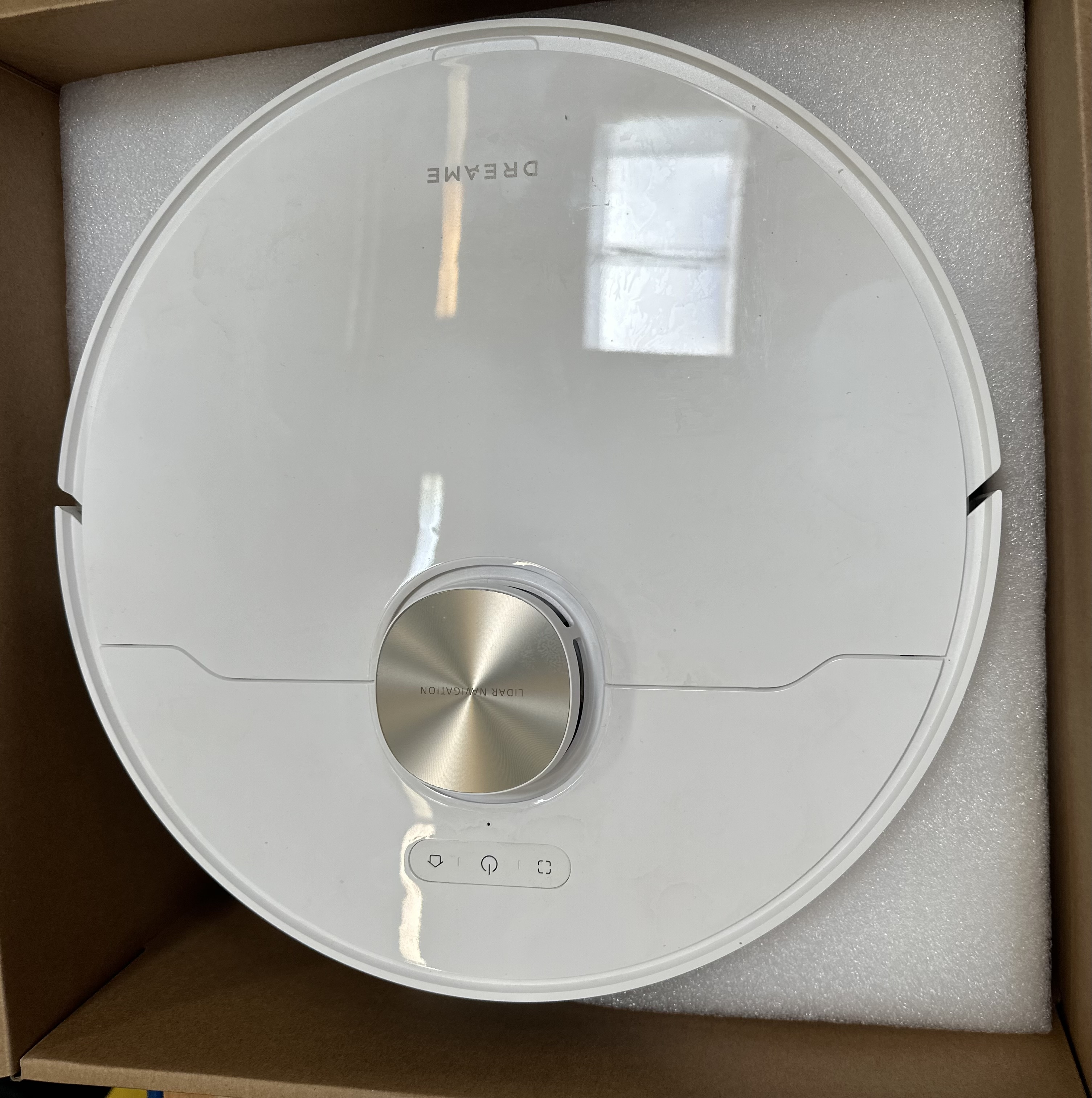 Biały odkurzacz robot Dreame wewnątrz kartonowego pudełka, widok z góry. Srebrne detale przycisku.