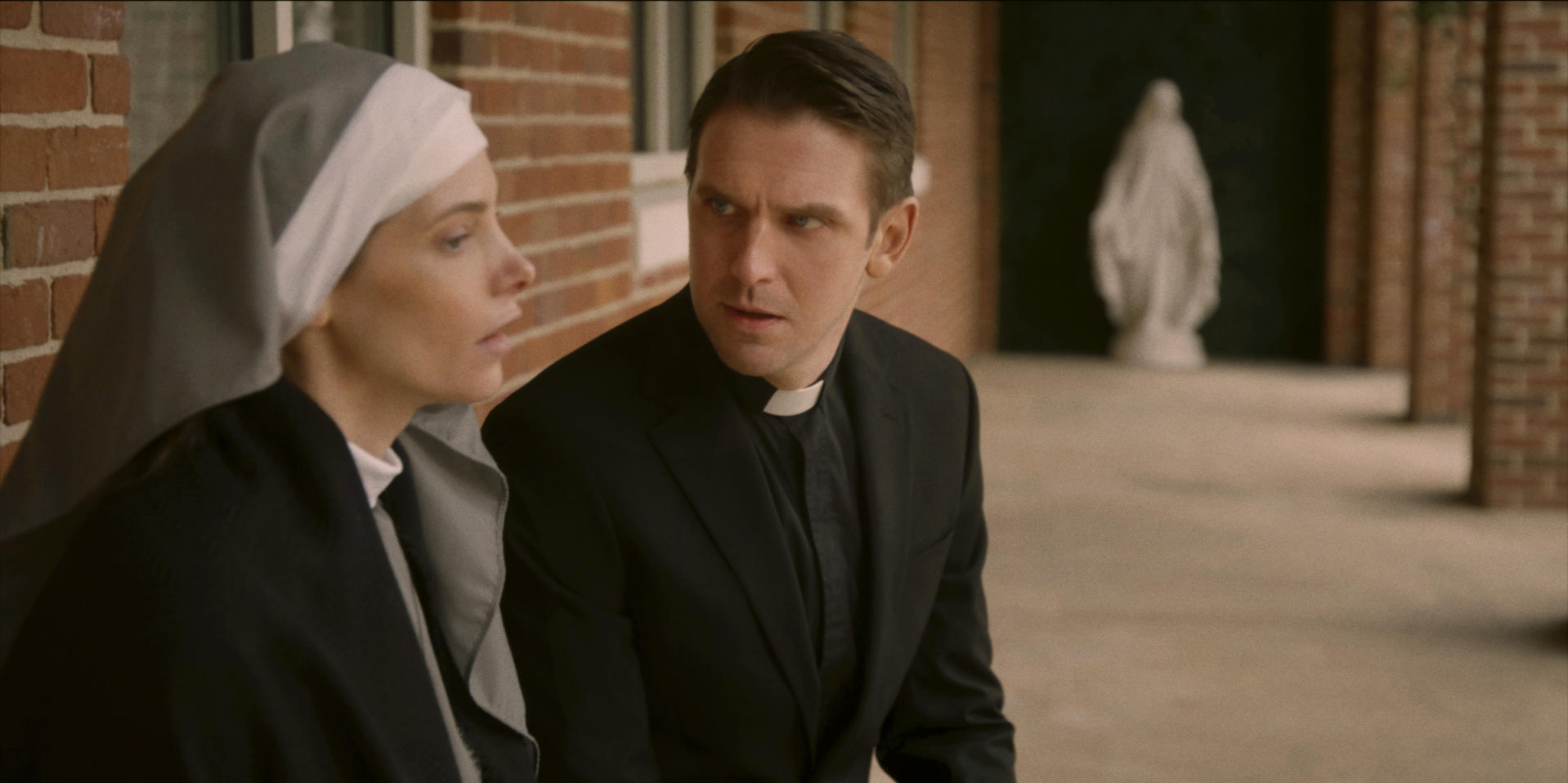 Eine Nonne und ein Priester unterhalten sich draußen, im Hintergrund eine Marienstatue.