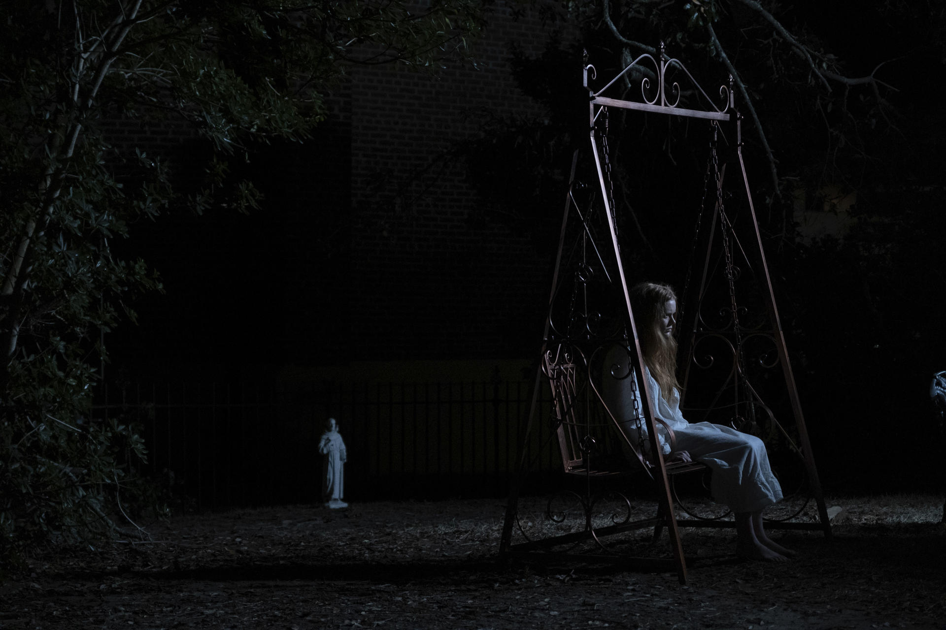 Eine Frau auf einer Schaukel in einem dunklen Garten bei Nacht, mit einer Statue im Hintergrund.