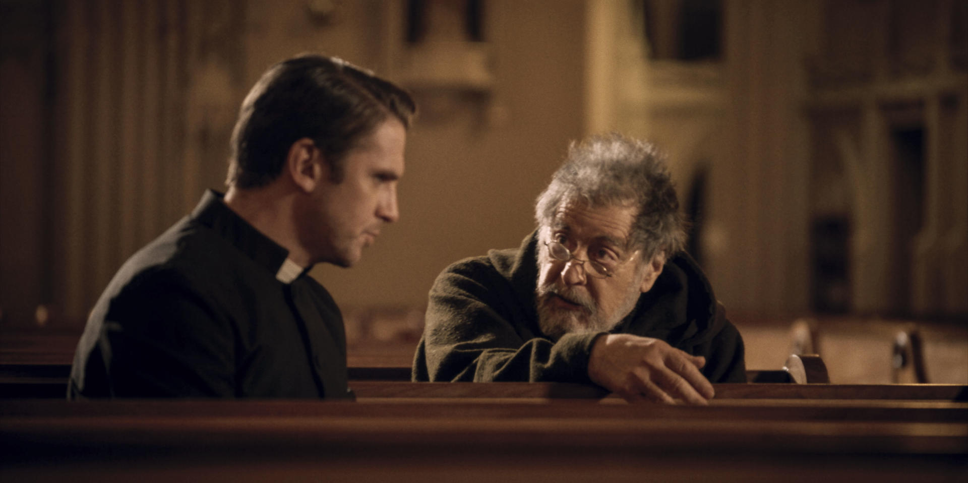 Zwei Männer sitzen in einer Kirche. Einer ist ein Priester, der andere scheint ein älterer Mann zu sein.