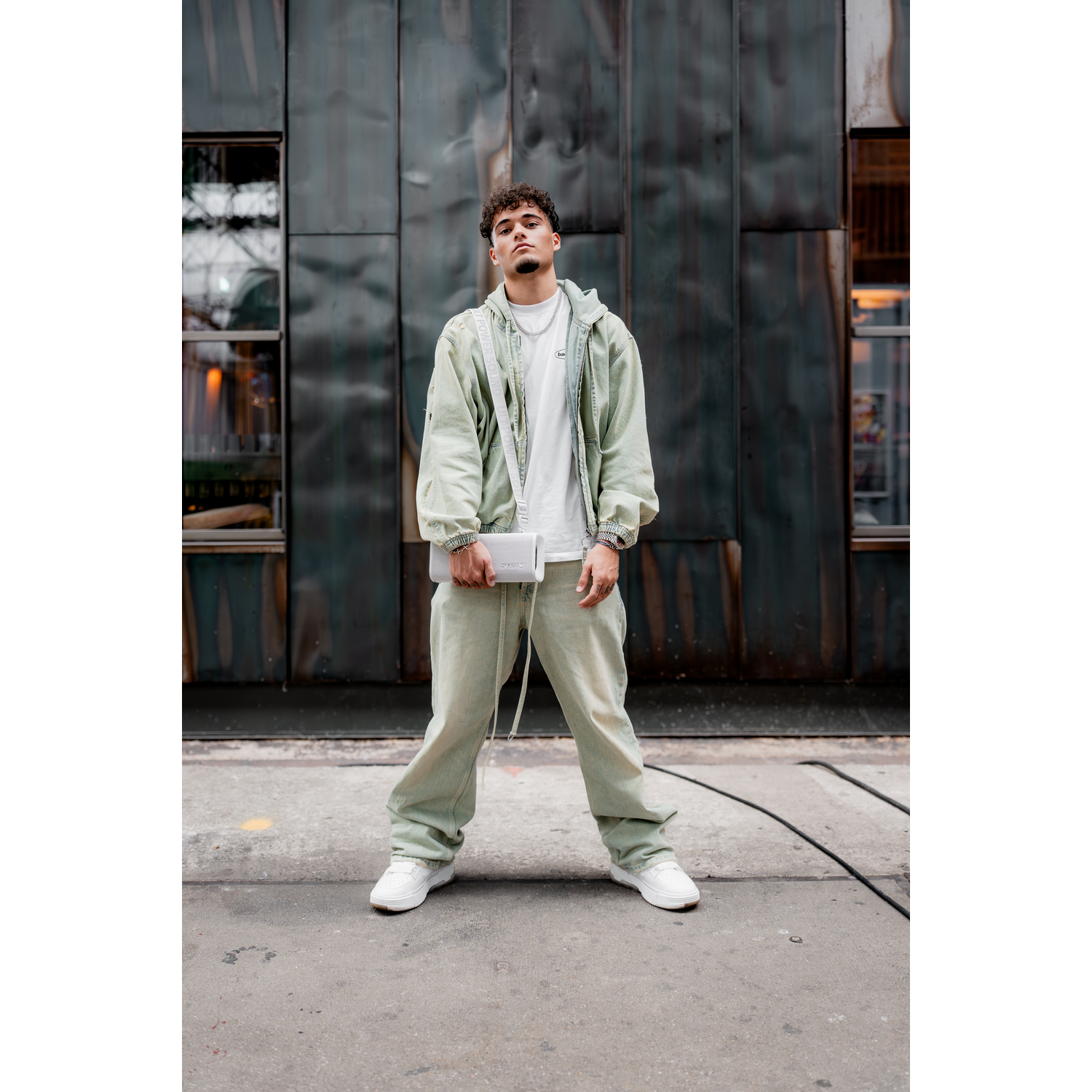 Een man in lichtgroene kleding staat voor een gestructureerde grijze metalen muur en houdt een kleine witte tas vast.