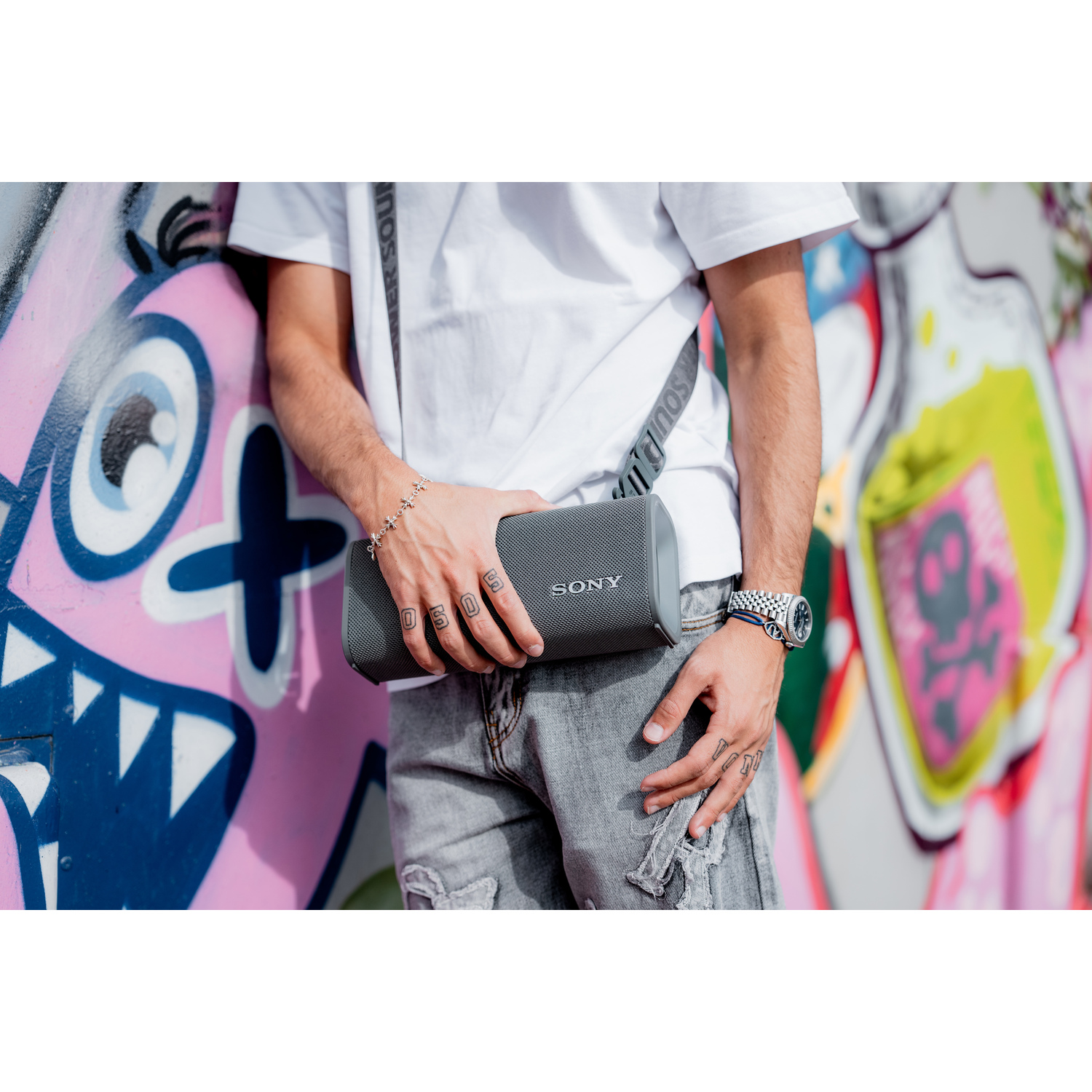 Een persoon houdt een grijze Sony-luidspreker tegen een graffiti-muur.