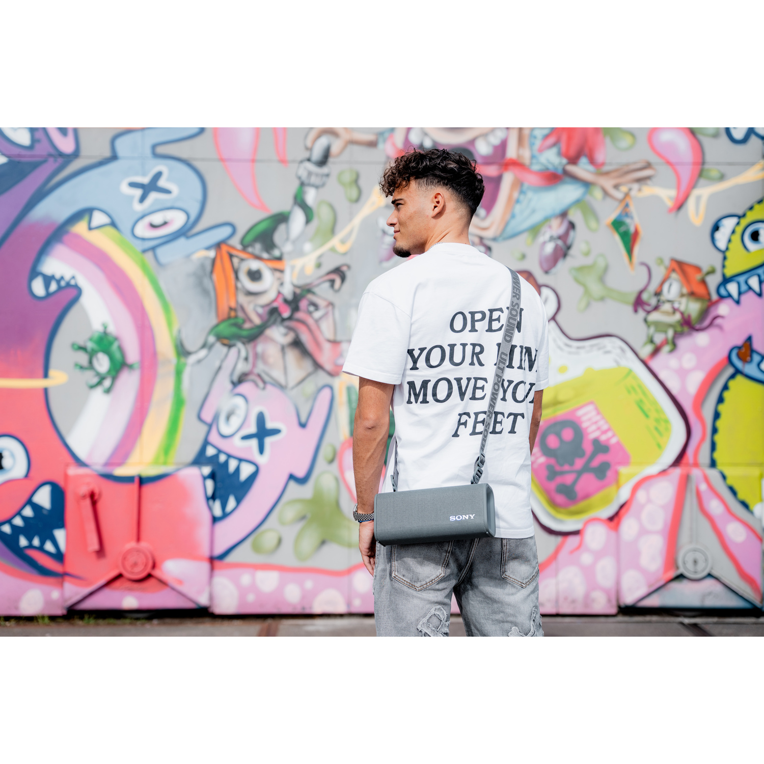 Jonge man met grijze luidspreker. Hij staat voor een graffiti muur. De luidspreker is grijs en heeft een riem.